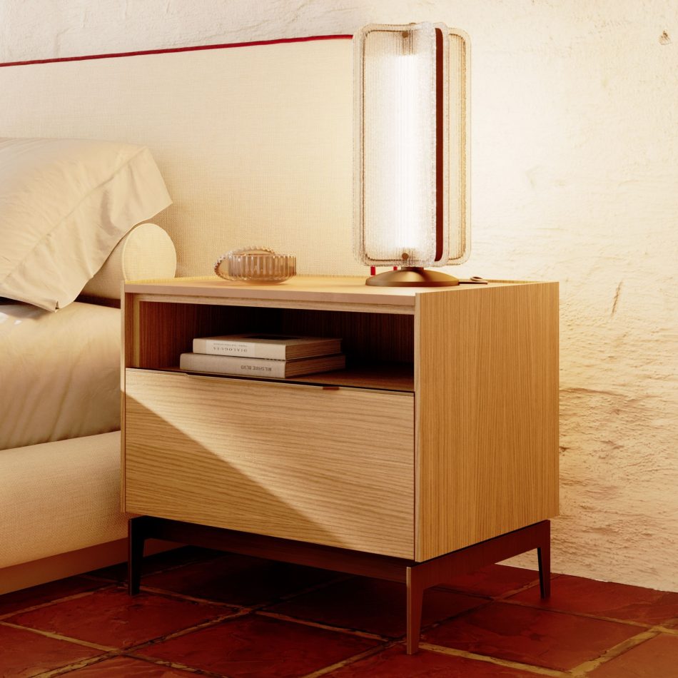 Warm, light-filled bedroom corner featuring the Landform Night Table I in natural oak wood texture, its open shelf holding a stack of books and the surface dressed with a glass object and sculptural table lamp. The finest wood grain and slender metal legs in a bronzed finish bring sophisticated bedroom furniture into a space defined by minimalist design and honest materiality.