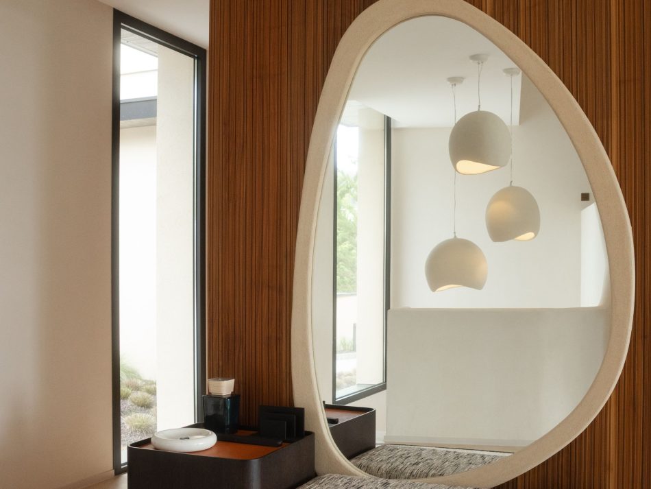 Close-up of the entrance hall's Stone Mirror, a organic-shaped piece with a cream stone frame set into the dark slatted solid wood wall. Its asymmetric teardrop silhouette reflects pendant lights and garden greenery beyond, softening the geometry of the space with a fluid, natural form. A defining moment of mirror design for wall and interior design inspiration, where a bespoke luxury console table below and wood design cladding create a quiet, considered composition that opens the home with intention.