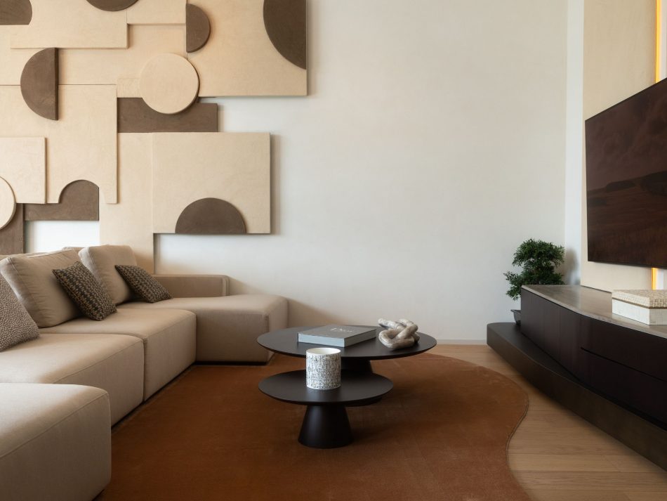 Living room detail showing the Landform Modular Sofa in cream furniture upholstery fabric alongside the Sahara Coffee Tables in matte black metal, their stacked organic geometries introducing sculptural depth to the seating area. The Stone TV Cabinet in dark wood design with polished brass accent lighting is visible to the right, completing a living room design where every piece earns its place through material honesty and quiet precision.