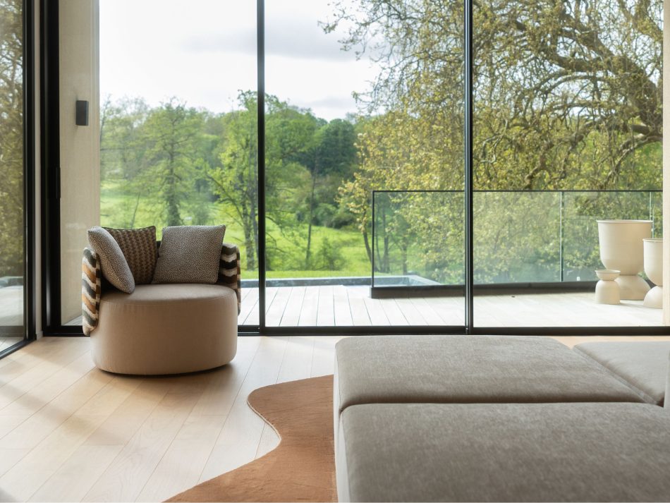 Floor-to-ceiling glazed living room opening onto a lush green landscape, with the Margem Armchair in warm taupe furniture upholstery fabric positioned by the window to take full advantage of the natural light and exterior view. The bespoke living room composition, with its organic-edged rug and modular sofa in the foreground, captures a moment of living room inspiration where luxury home furniture and nature exist in quiet dialogue.