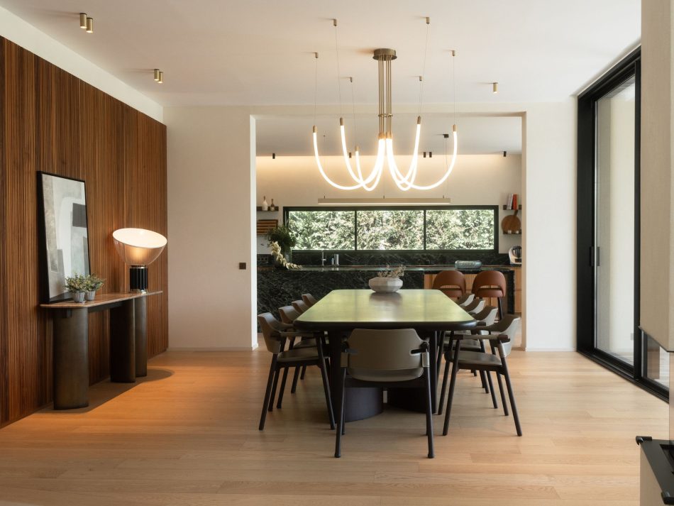 Open-plan dining room featuring the Dolomites Dining Table in dark wood design surrounded by upholstered dining chairs in warm brown tones, beneath a sculptural branching chandelier with gold-plated brass fittings. The Dolomites Console sits against the dark slatted solid wood feature wall to the left, styled with a table lamp and artwork, while a marquina marble kitchen backsplash frames the garden view beyond. A complete and considered luxury dining room furniture setting where materiality and light define the atmosphere.