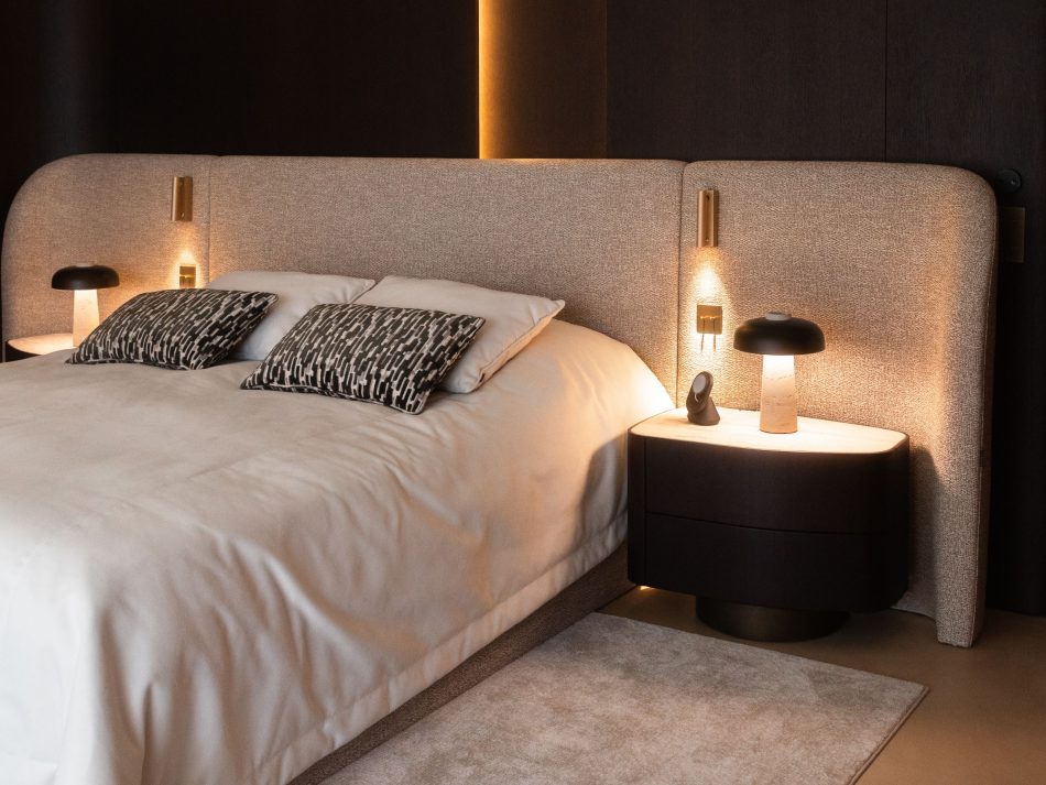 Master suite detail featuring the Stone Bed with a generously proportioned sculptural headboard in warm greige furniture upholstery fabric, flanked by Stone Night Table II pieces in dark wood design with rounded forms and ambient table lamps. A deeply considered luxury master bedroom furniture composition where gold-plated brass wall sconces and layered indirect lighting create an atmosphere of immediate calm.