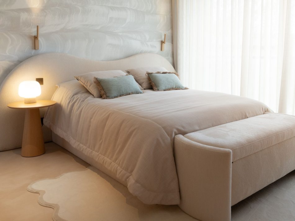 Children's bedroom featuring a sculptural bed with a deeply curved organic bouclé furniture upholstery fabric headboard in soft cream, dressed with sage green fringed cushions and a matching bench at the foot. The Olivier Side Table in natural solid wood with a conical base sits beside the bed with a glowing table lamp, while gold-plated brass wall sconces and sheer curtains complete a bespoke bedroom defined by lightness and calm.