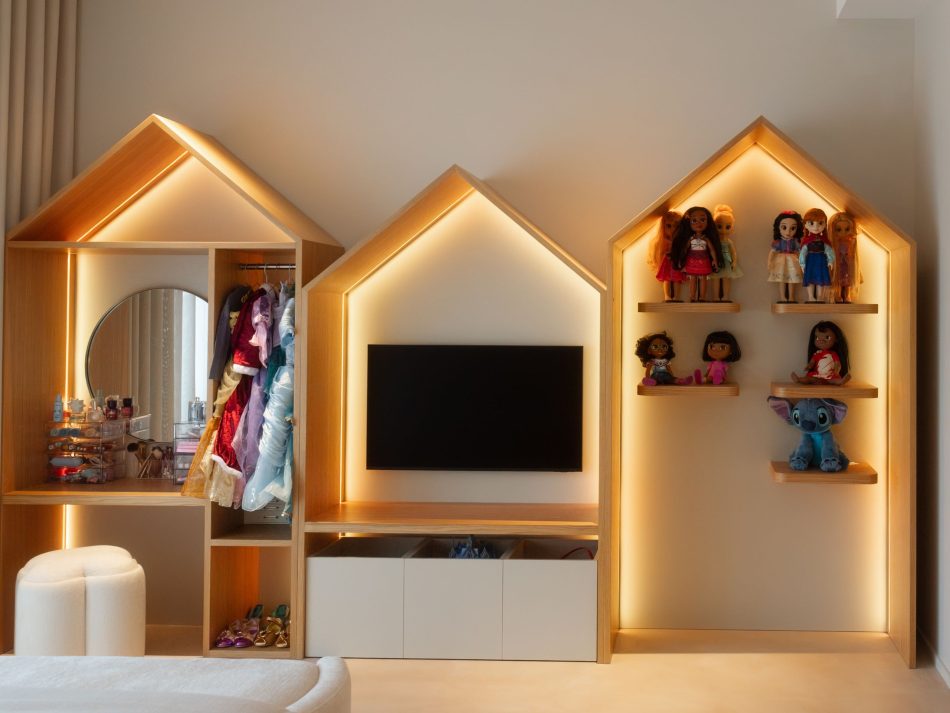 Children's bedroom featuring a bespoke wall composition of three house-shaped joinery units in natural wood texture, each backlit in warm amber light. The left unit houses a dressing area with a round mirror and costume rail, the central unit frames a television with white lacquer storage below, and the right unit functions as an open decorative bookcase with floating shelves displaying toys and figures. A bespoke bedroom solution where handmade furniture craft and imaginative design come together in a space genuinely made for childhood.