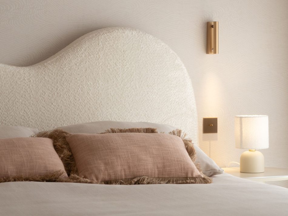 Child's bedroom detail featuring the sculptural wave-form bouclé furniture upholstery fabric headboard in soft white, dressed with blush pink fringed cushions and a warm patterned pillow. A gold-plated brass wall sconce casts warm directional light beside a compact ceramic table lamp, completing a bespoke bedroom composition where organic forms and soft natural materials create a room that feels light, calm, and genuinely made for childhood.