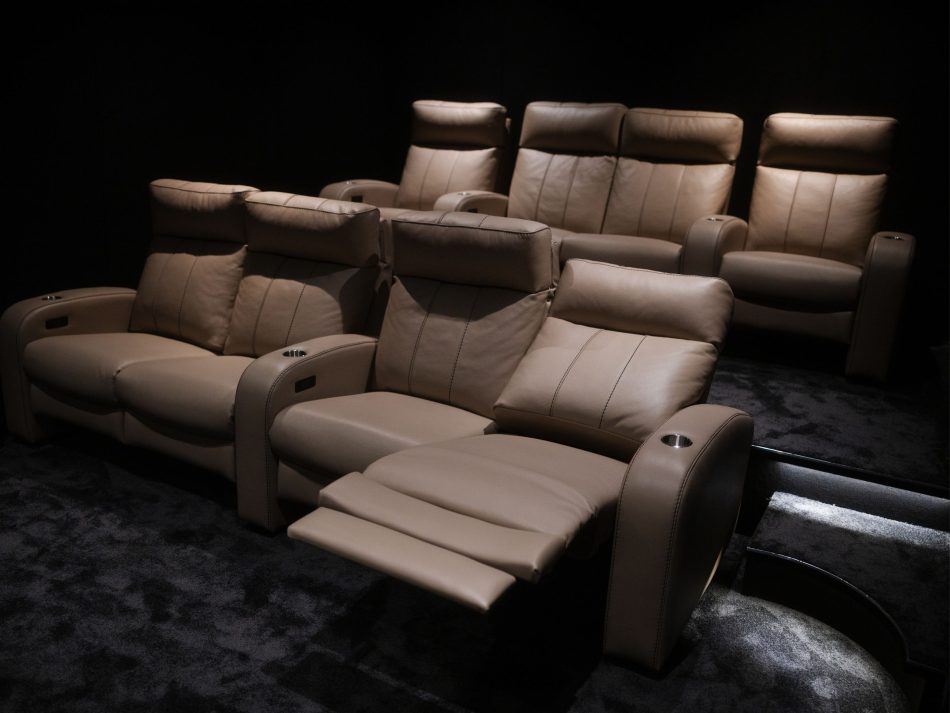Private home cinema featuring two rows of Lumière Cinema Armchairs in warm taupe leather upholstery, one with the footrest fully extended, arranged on a dark carpet against a black acoustic backdrop. Integrated cup holders and stainless steel cup inserts in the armrests complete the experience, creating an atmosphere of high end furniture comfort and immersive interior design inspiration for a truly private cinema setting.
