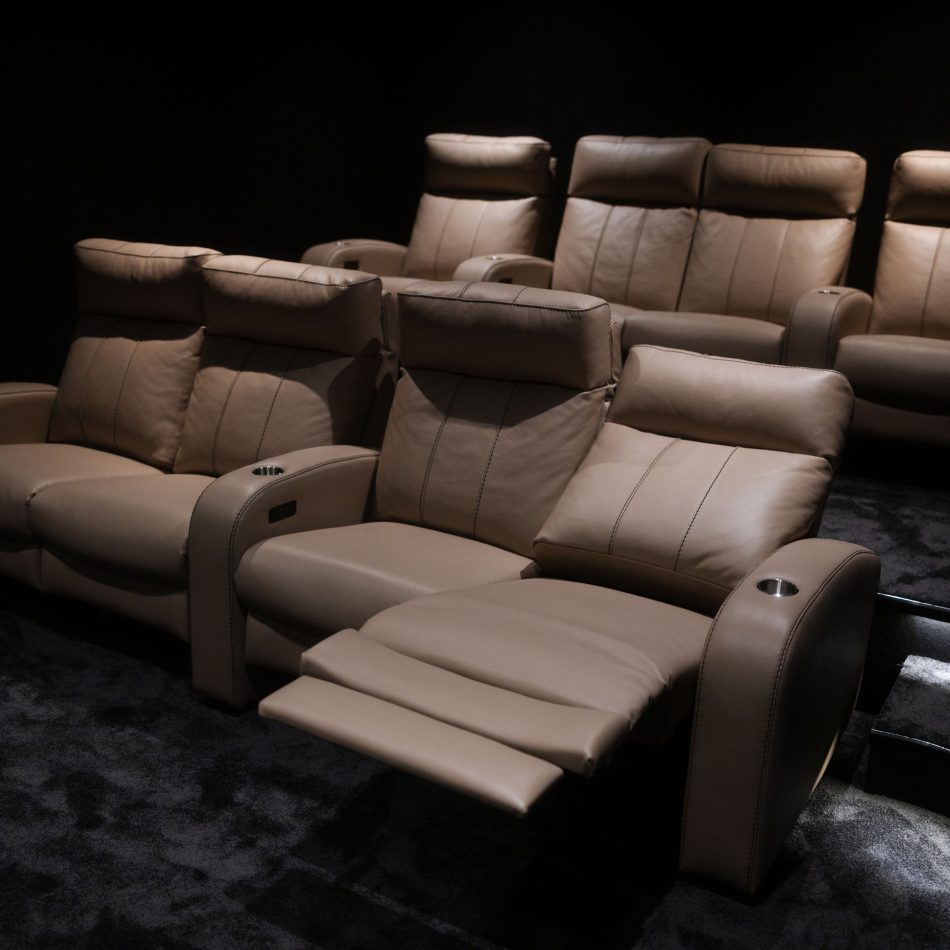 Lumière Cinema Armchair arranged in rows within a private cinema room, upholstered in warm taupe leather upholstery with contrast topstitching, integrated cup holders, and one seat shown with footrest extended. A luxurious furniture installation that brings hospitality furniture standards into the private home, where every seat is conceived for immersive, extended viewing comfort.