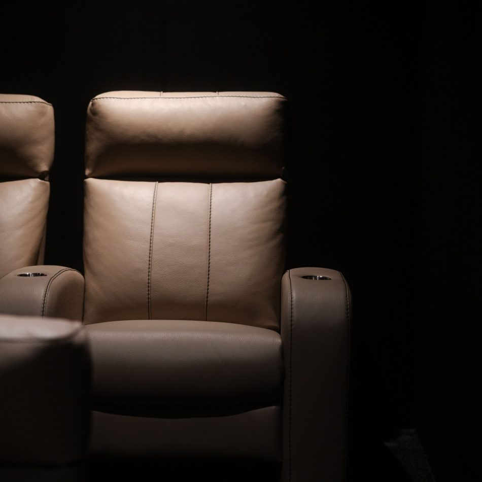 Close front view of the Lumière Cinema Armchair backrest and headrest in warm taupe leather upholstery, dramatically lit against a dark cinema interior, with integrated cup holders visible on both armrests. The majestic and sleek volume of the backrest and the refined stitching detail position this as a piece of high end luxury furniture designed to elevate the private cinema experience.