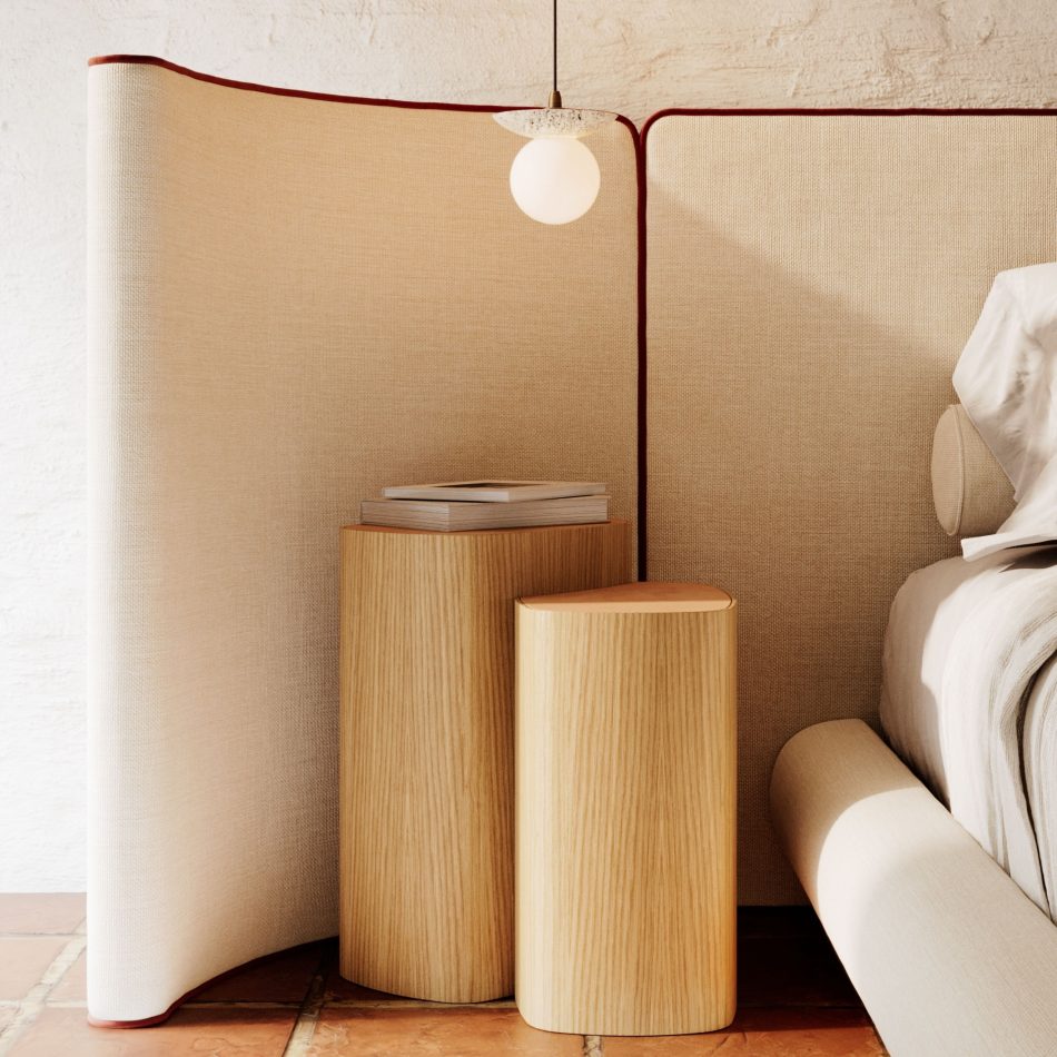 Close-up bedroom corner with the Natur Bed's curved asymmetric headboard panels in natural greige furniture upholstery fabric with terracotta piping, framing two cylindrical solid wood side tables of varying heights stacked with books beneath a globe pendant lamp. The wood texture of the tables and the soft textile of the headboard create a moment of duality and contrast that defines this sophisticated bedroom furniture composition at its most intimate.