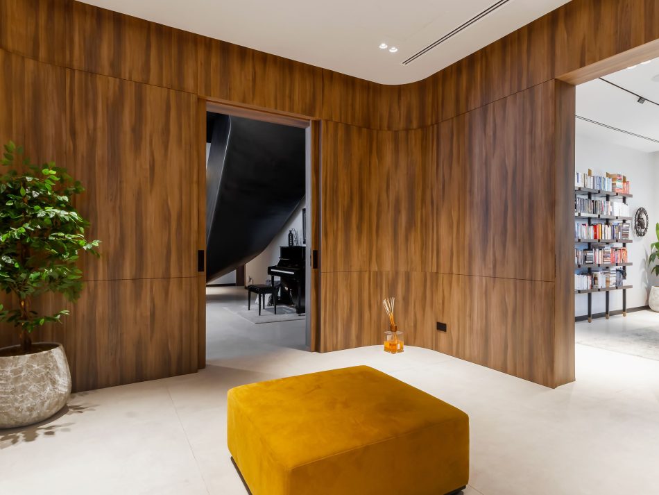 A wide hallway wrapped in warm wood texture panelling opens onto a music room beyond, where a grand piano anchors the space. A decorative bookcase lines the right wall, filled with books and objects. At the centre, the Landform Puff in mustard velvet furniture grounds the transition between rooms. Bespoke furniture and architectural restraint define this residence, where every detail is built into the space rather than added to it.