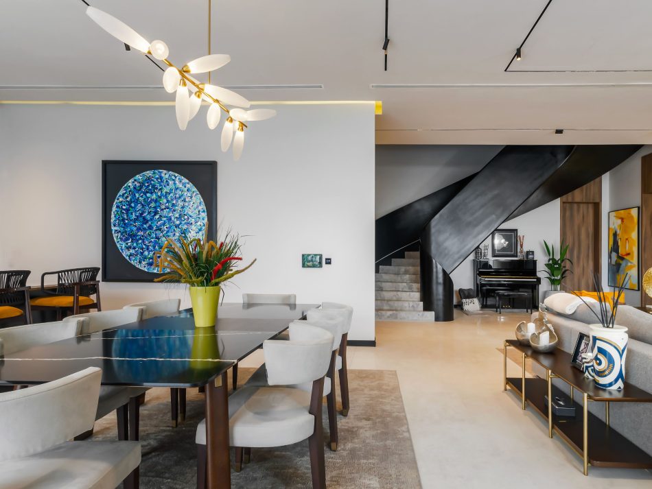 An open-plan residence where the Landform Dining Table and Her Chair anchor the bespoke dining room beneath a sculptural brass chandelier. A large circular painting in deep blues commands the wall, while a dramatic black staircase draws the eye through the full depth of the space. Luxury dining room furniture and architectural scale define a home where every element holds its own.