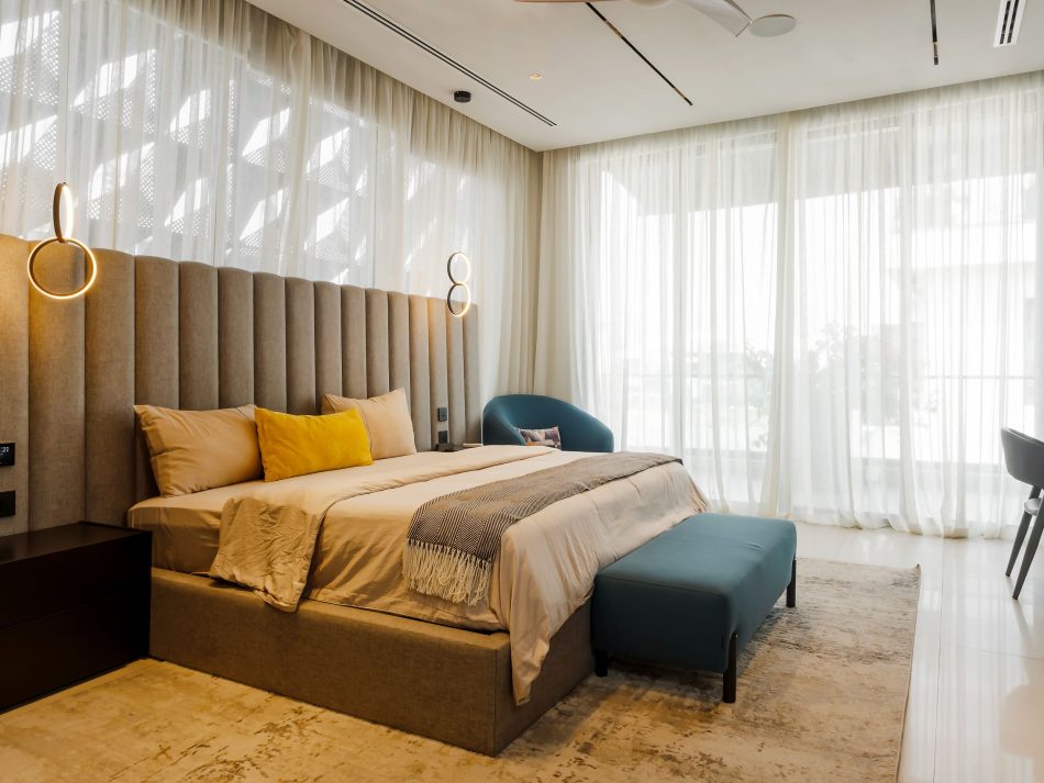 A light-filled master suite centred on the Milan Bed, its floor-to-ceiling channelled headboard in warm grey upholstery softened by sheer curtains and natural light. The Sevilha Night Tables in dark wood create duality and contrast, while the petrol blue Club Armchair and Terra Bench bring intentional colour to an interior defined by luxury master bedroom furniture.