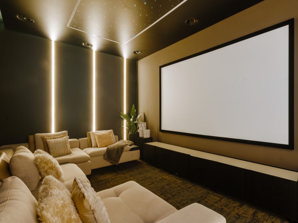 A home cinema where the Margem Sofa in cream upholstery faces a full-width projection screen, surrounded by dark walls and vertical amber lighting strips. The Sevilha tv cabinet beneath the screen keeps the composition grounded with quiet precision. A star-lit ceiling completes the atmosphere, showing how bespoke furniture can turn a technical room into something deeply enveloping.