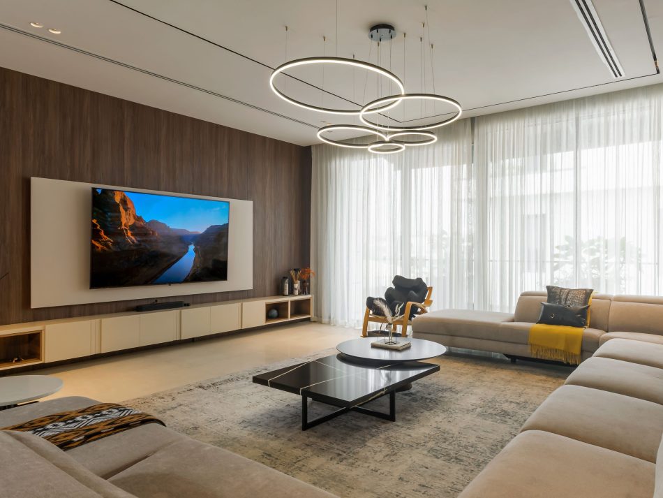 A secondary living room where a modular sofa in cream upholstery wraps around the Sahara Coffee Table in dark marquina and the Slender Coffee Table in a lighter finish. The Sevilha tv cabinet extends across the timber-panelled wall, combining closed storage with open display niches. A cluster of circular pendant lights overhead reinforces the living room architecture, drawing the eye through a space designed for a slower, more private kind of comfort.