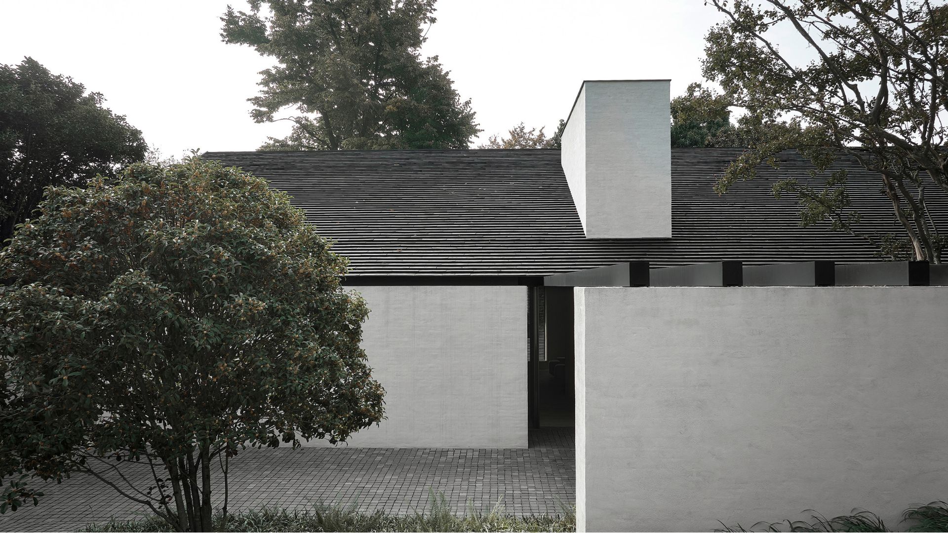 A residence by Vincent Van Duysen, the B Residence, showcases a low-pitched dark timber roof against white walls. with a tall chimney volume rising above the roofline. Dense garden greenery frames the structure, while a sheltered walkway beneath the eaves leads toward the entrance. The composition reflects an "architecture first" sensibility built on minimalist design, harsh and strict lines, and a quiet, monumental presence rather than ornament.