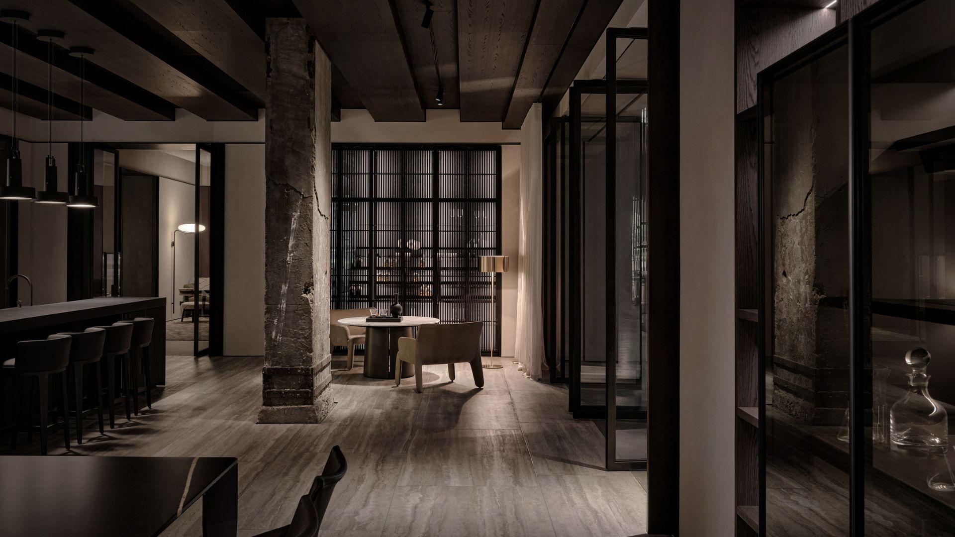A dimly lit interior at the Molteni&C Shanghai Showroom reveals an exposed concrete column among rich wood texture ceiling beams and travertine flooring. A rounded dining table in dark brass sits beneath a sculptural floor lamp, framed by a slatted timber screen. Glass-fronted stainless steel cabinets line the wall, embodying Van Duysen's noble simplicity and harsh and strict lines.
