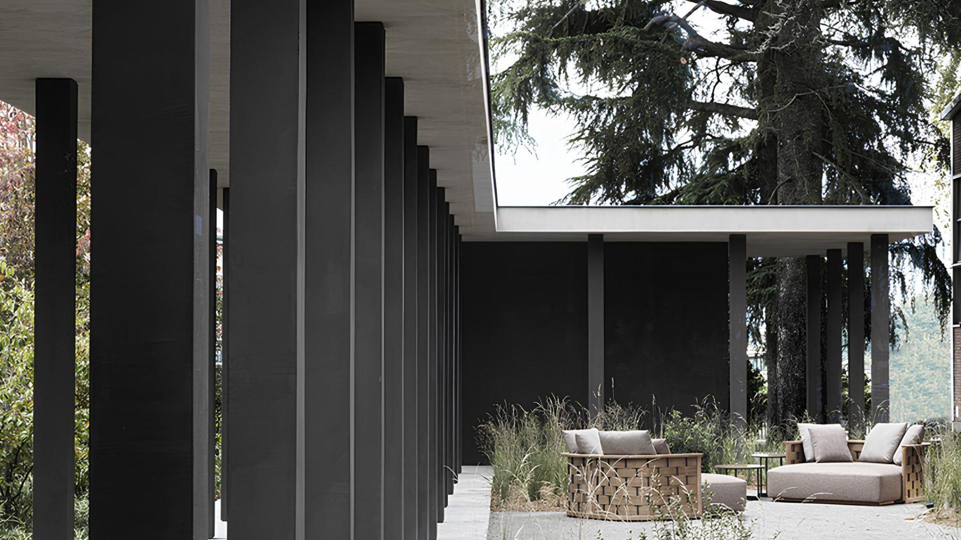 A repeating colonnade of dark stainless steel columns lines a covered walkway, framing a view of mature trees and a garden seating area beyond. Woven rattan and pale upholstery fabric furniture sits among tall grasses on a stone terrace, while a cantilevered concrete roof extends overhead. The composition reflects Van Duysen's material discipline, where harsh and strict lines and restrained colour let texture and light define the atmosphere.