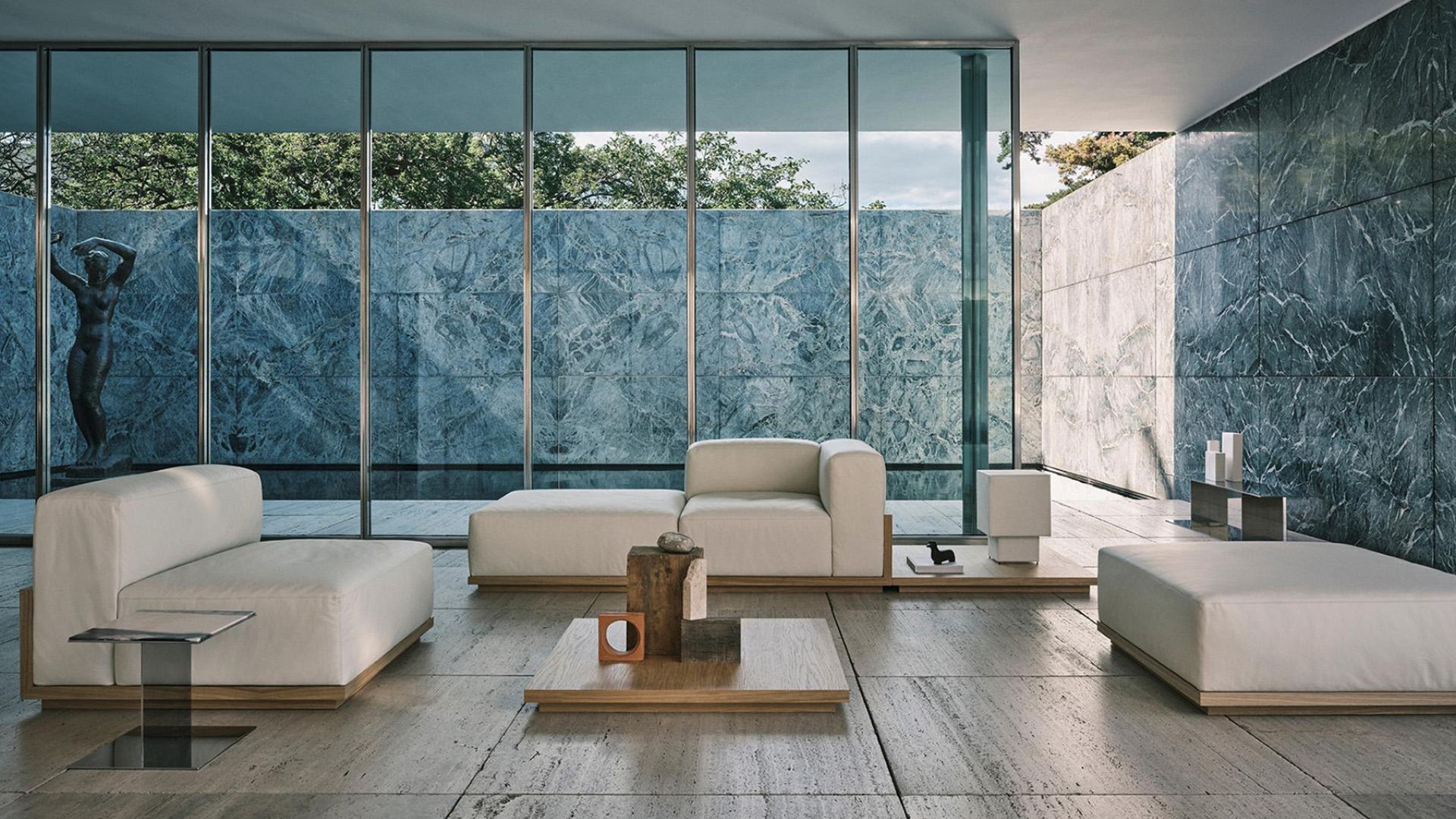 From Zara Home's Collection 04 by Vincent Van Duysen, a low-slung sofa in cream upholstery fabric sits on a travertine floor, paired with a coffee table in pale wood texture holding sculptural ceramic objects. Floor-to-ceiling glazing frames a courtyard lined with veined marble walls and a classical bronze statue. The arrangement reflects Van Duysen's architectural approach to furniture, where pieces create spatial relations rather than simply fill the room.