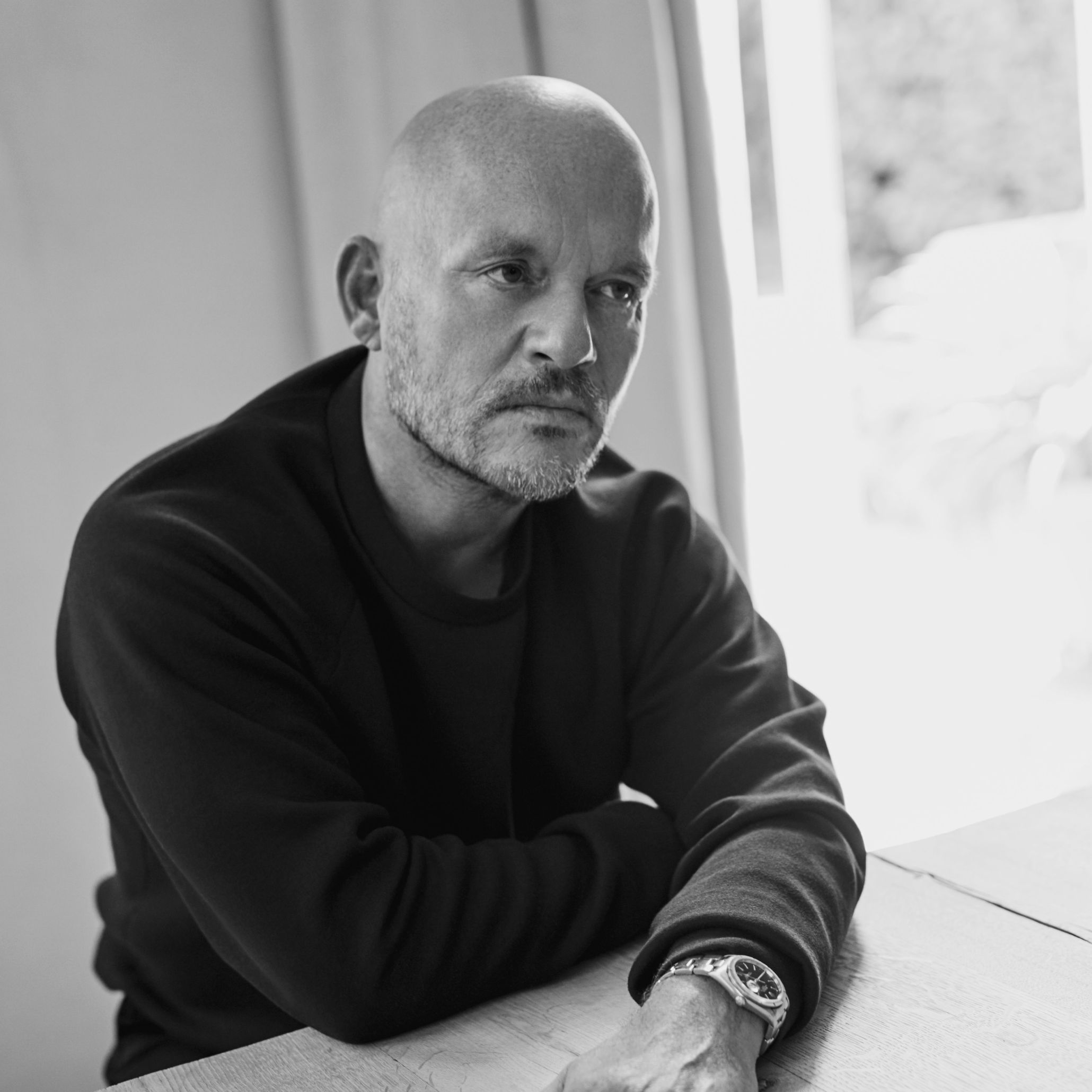 Black and white portrait of Vincent Van Duysen, founder of Vincent Van Duysen Architects, seated with arms crossed beside a window.