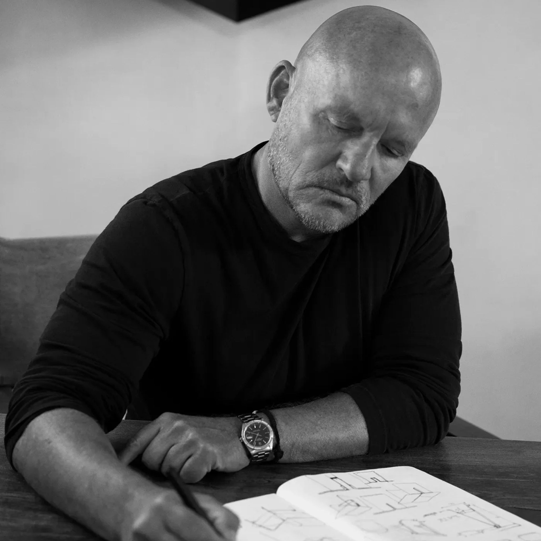 Black and white portrait of Vincent Van Duysen reviewing architectural sketches at a wooden table.