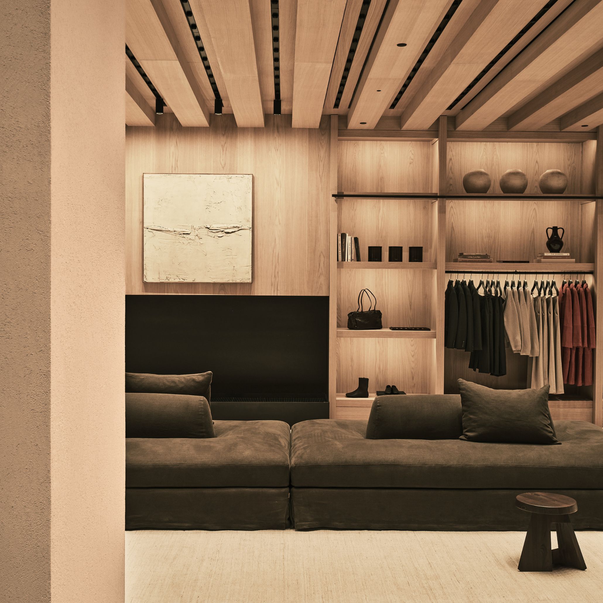 Inside Zara Diagonal in Barcelona, a deep brown modular sofa in leather upholstery sits before a fitted wood texture display wall holding garments, accessories and ceramic objects with quiet precision. A textured plaster artwork hangs above a built-in fireplace, while a sculptural dark wood stool grounds the foot of the seating. Light filters across the timber surfaces, illustrating Van Duysen's material honesty and human scale.
