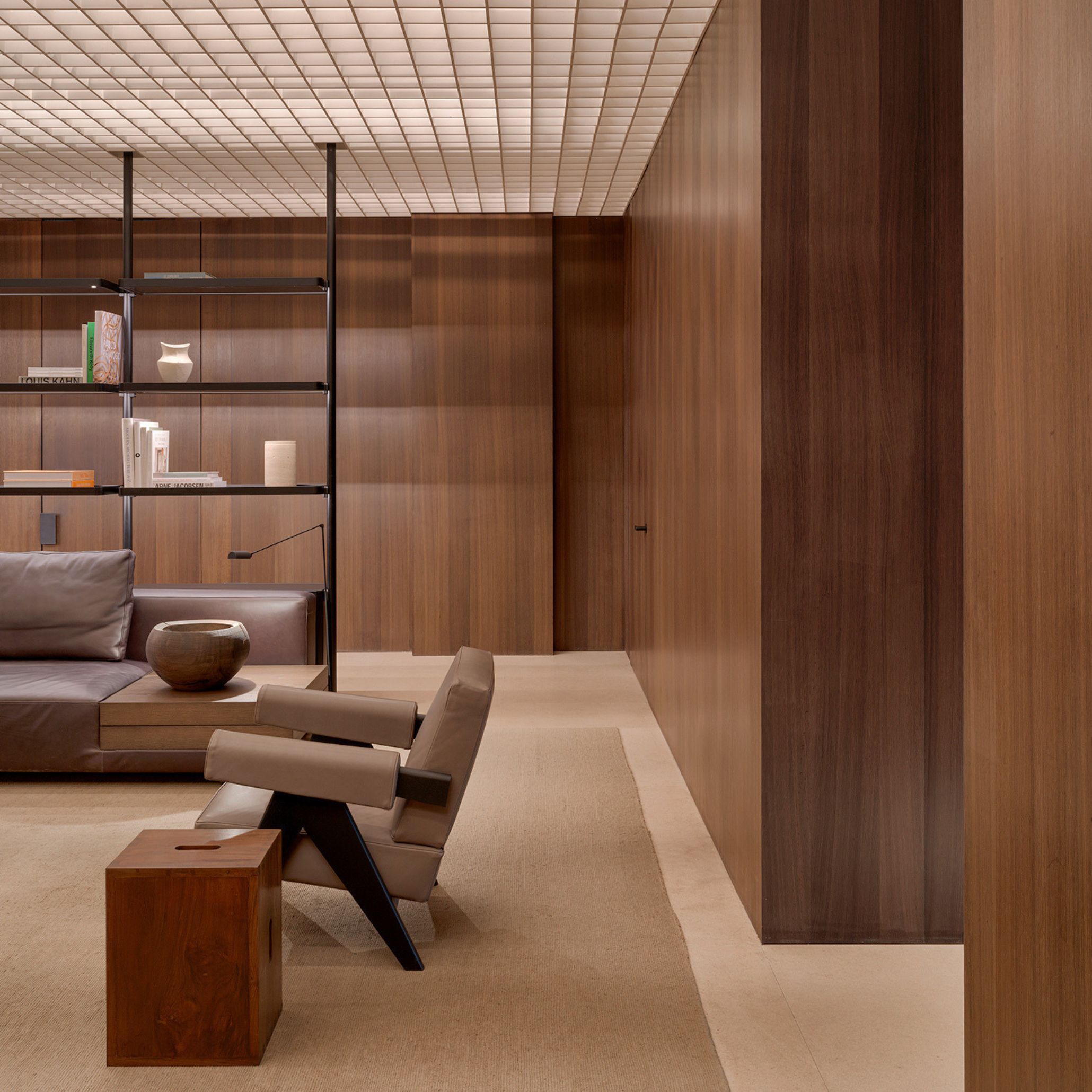 At M Offices in New Delhi, a leather sofa in deep aubergine sits beneath an open black steel decorative bookcase holding books and ceramic vessels, set against full-height wood texture panelling. A pair of armchairs in taupe leather upholstery with angular dark frames face the seating, beside a solid wood cube stool. A coffered ceiling in pale tile catches the light, reinforcing Van Duysen's restrained, material-led language.