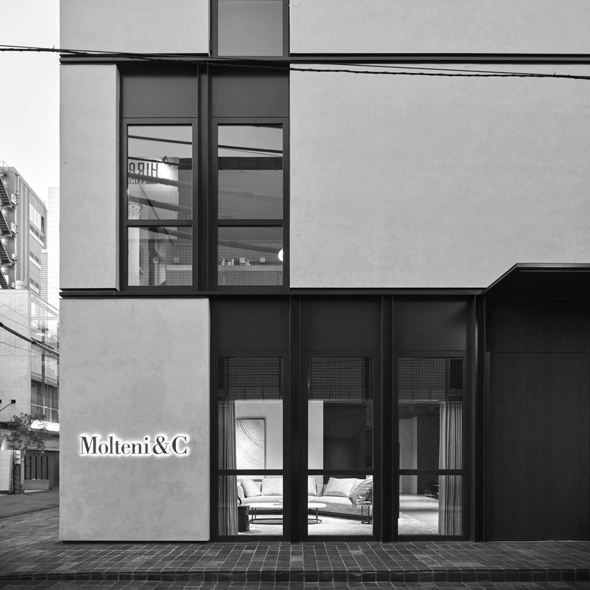 The Palazzo Molteni Tokyo façade in Minami Aoyama, with stainless steel framed glazing set within rendered concrete volumes. Illuminated signage marks the entrance, while a glimpse of upholstered seating is visible through the storefront windows below. Solid masses and full-height openings create a quiet, geometric rhythm informed by Japanese architectural heritage.