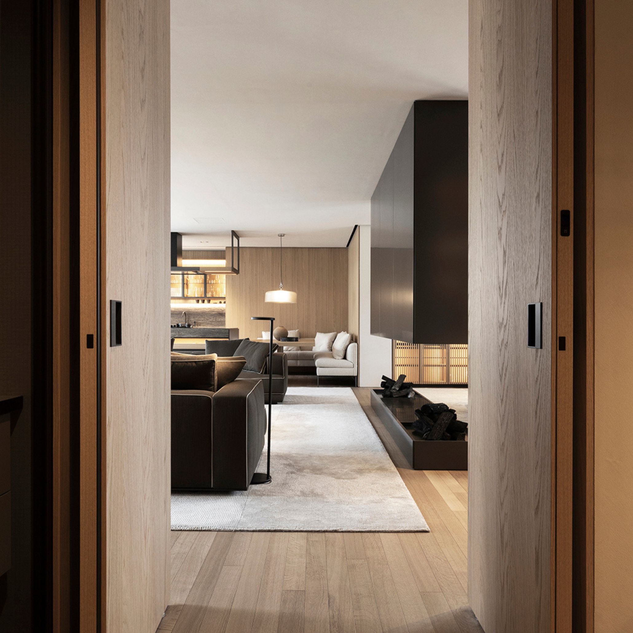 A view through sliding wood texture doors into the Palazzo Molteni Tokyo interior, where dark leather upholstery seating faces a sunken fireplace lined with stacked firewood. Warm timber flooring and panelling extend toward the kitchen beyond, framed by a sculptural black volume suspended above the hearth. The composition reflects a quiet dialogue between European design culture and Japanese restraint.