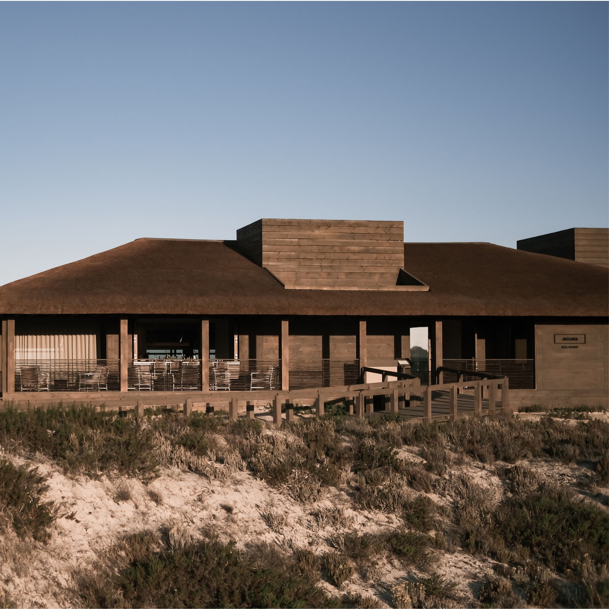 The JNcQUOI Beach Club rises from coastal dunes in Comporta, its thatched roof and wood texture cladding echoing the vernacular of local fishermen's huts. A sheltered terrace lined with rattan furniture runs beneath the wide eaves, while a tall chimney volume in stained timber breaks the roofline. Set against a clear blue sky, the structure stays low and absorbed into the landscape, true to Van Duysen's site-specific restraint.