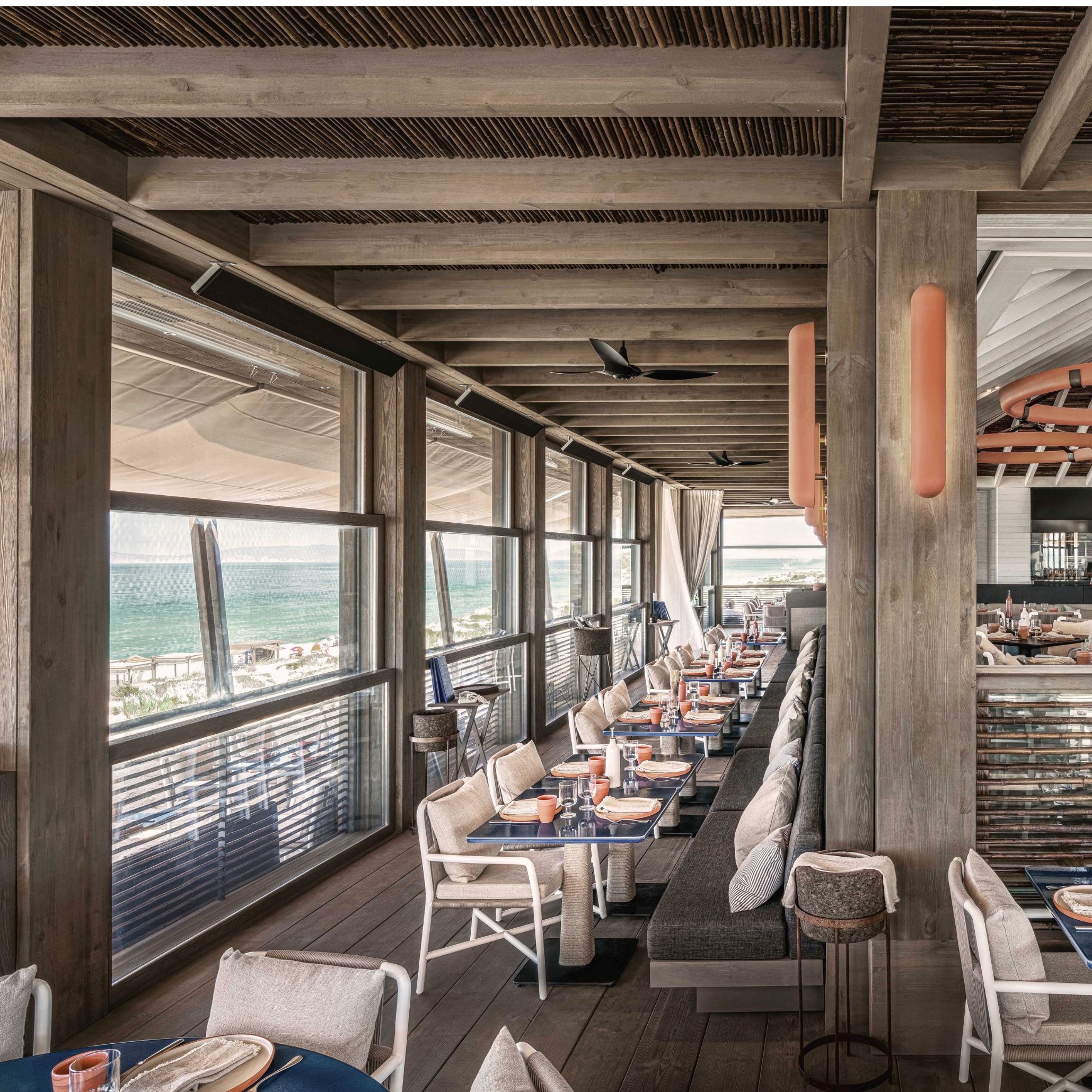 Inside the JNcQUOI Beach Club, a long banquette in grey upholstery fabric runs beneath exposed caned ceilings and timber beams, facing full-height windows that open onto the Atlantic. Terracotta table settings and woven chairs in pale linen bring warmth to the dining terrace, while coral-toned wall sconces add a coastal accent. The space reflects Van Duysen's deliberate contrast between quiet exterior and animated interior character.