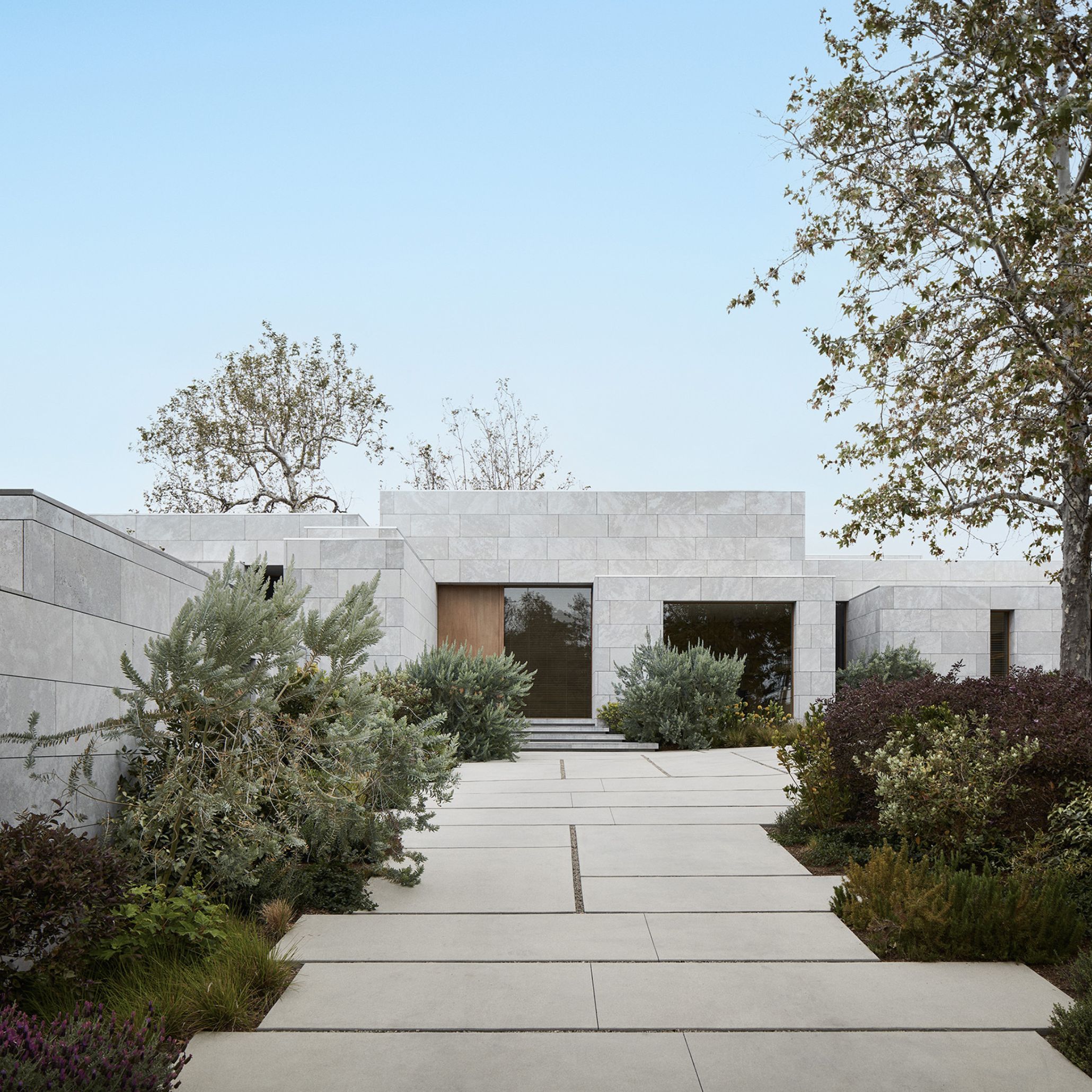 The AN Residence in Los Angeles presents a monolithic stone volume, its facade clad in pale marble blocks set in stacked, offset geometries. A wood-framed entrance door sits beneath a sculptural overhang, approached by a concrete path lined with native drought-tolerant planting. Mature trees rise behind the rooflines, reinforcing the residence's quiet integration into its secluded plateau setting.