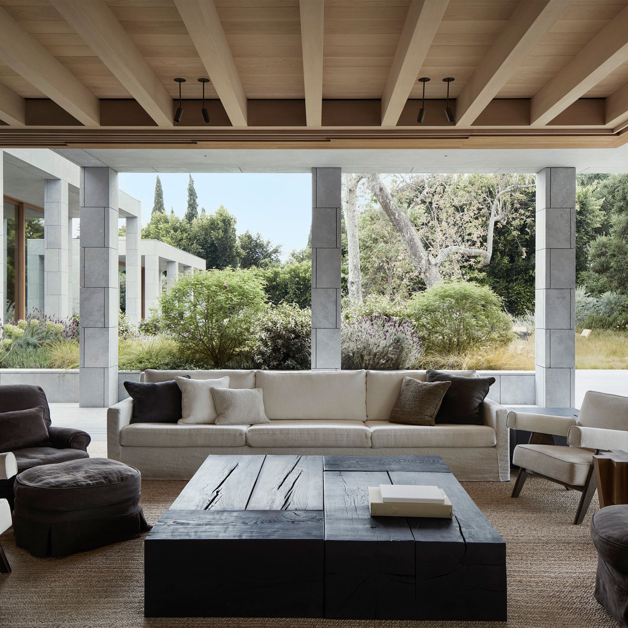 A covered terrace at the AN Residence frames the surrounding garden through deep stone columns, with a cream upholstery fabric sofa and dark linen armchairs arranged around a sculptural blackened wood texture coffee table. The timber-beamed ceiling extends the indoor language outward, dissolving the boundary between inside and out. Mature trees and soft planting complete a setting built on the city's mid-century Case Study tradition.