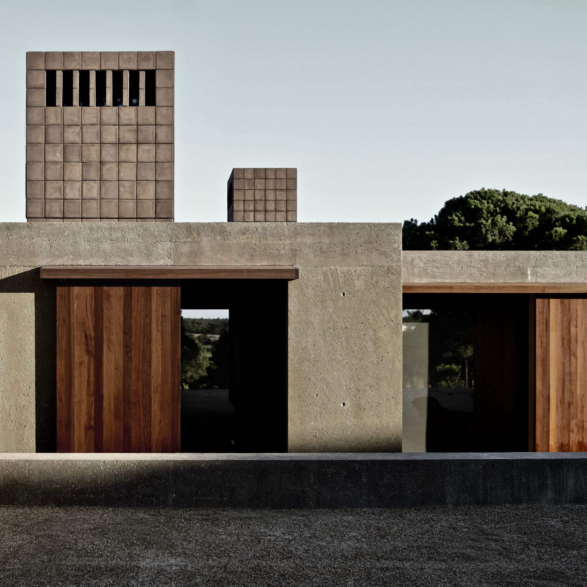 Casa M's exterior in Melides reveals two sculptural chimney volumes clad in pale square tile, rising above bone-coloured exposed aggregate concrete walls. Sliding doors in warm vertical finest wood panelling open onto dark interior thresholds, framed by the raw, pitted texture of the concrete surface. Mature pine trees rise beyond the rooftop, reinforcing the structure's quiet integration into its sandy Alentejo surroundings.