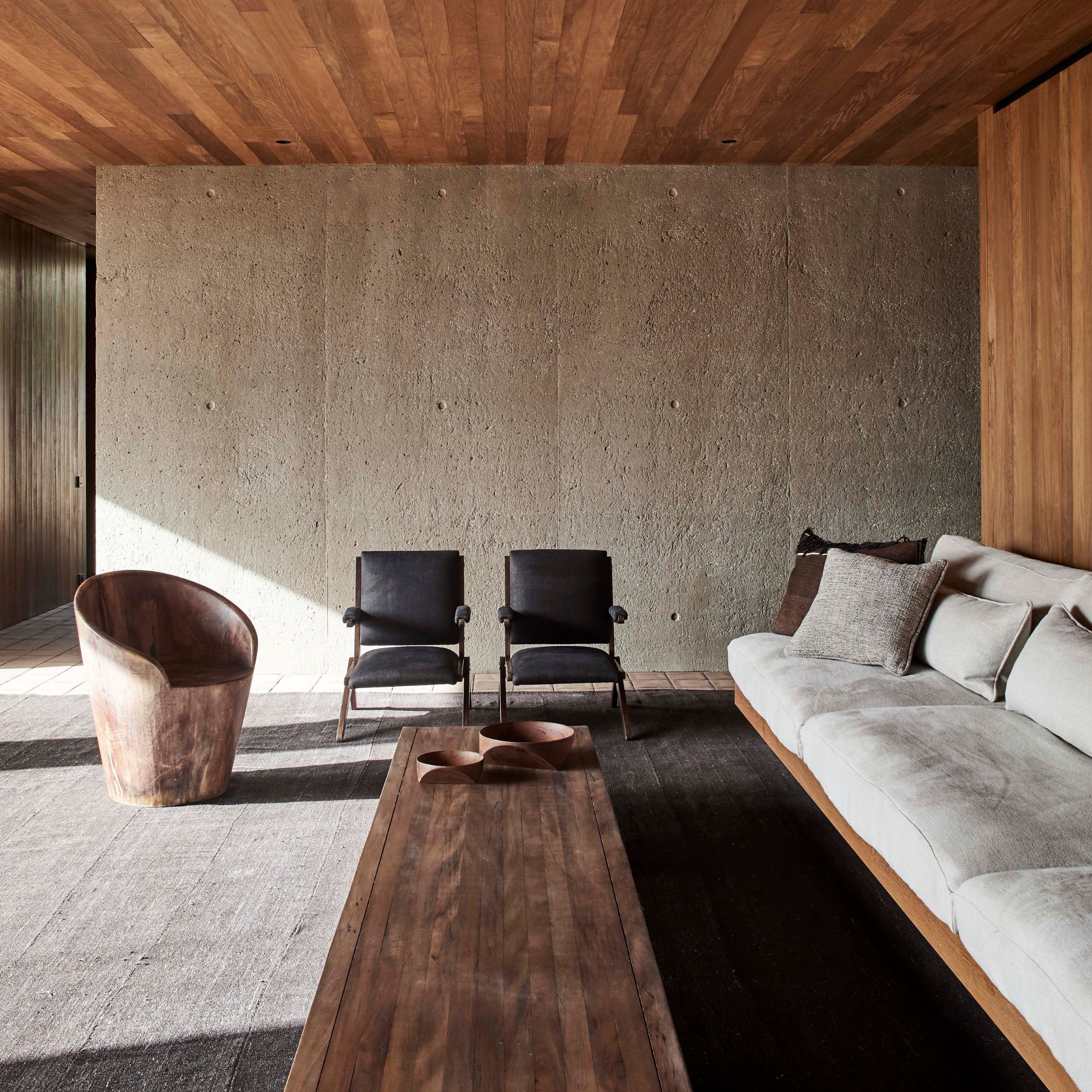 Inside Casa M, a low sofa in pale white upholstery fabric sits beneath a warm vertical wood texture ceiling, facing a long solid wood coffee table holding two carved bowls. Two black leather-framed armchairs and a curved tub chair in aged finest wood complete the seating, set against a raw, board-marked concrete wall. Shifting natural light traces the texture of the wall, embodying the house's non-ornamental attitude.