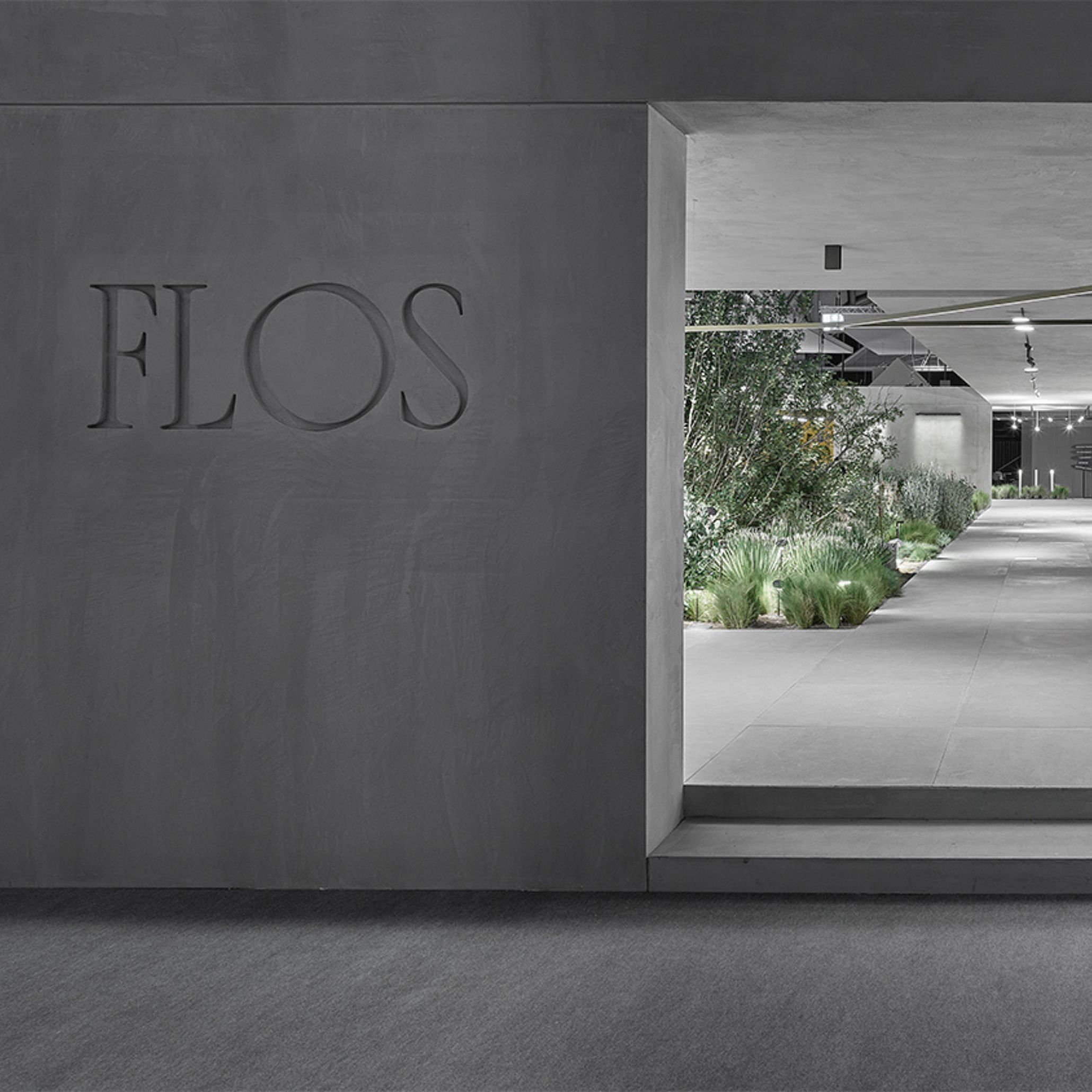 The Flos logo is carved into a textured grey concrete wall at a fair stand, opening onto a softly lit corridor lined with greenery and exhibition spaces beyond. The minimal signage and raw plaster surface reflect Van Duysen's minimalist design language, part of his longstanding lighting design collaboration with the brand.