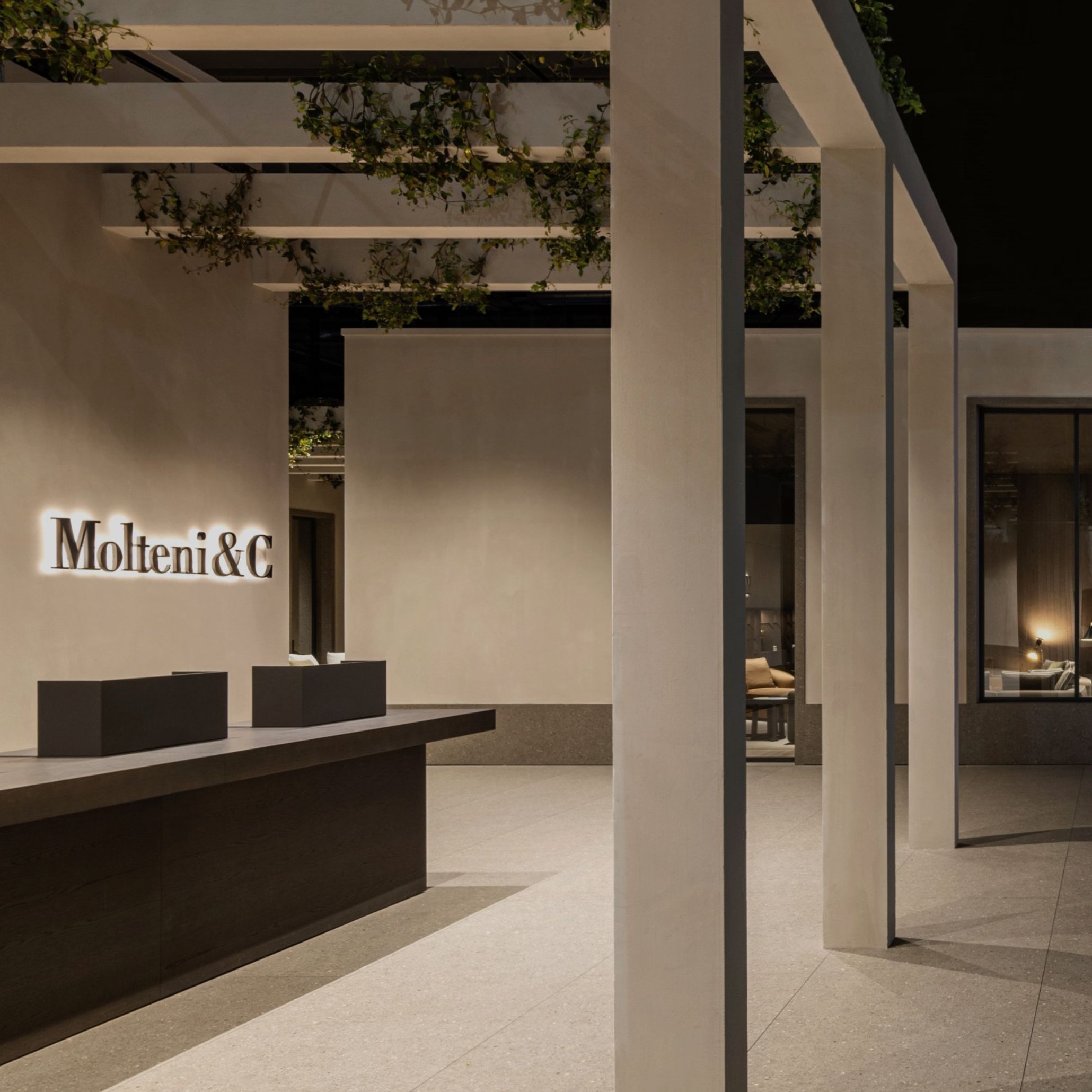 The Molteni&C signage glows softly against a plaster wall within a pergola structure, its slim stainless steel columns supporting overhead beams draped in trailing greenery. A dark stone reception counter with harsh and strict lines sits to the left, with furnished interiors visible through doorways beyond, reflecting Van Duysen's Molteni&C collaboration as Creative Director.