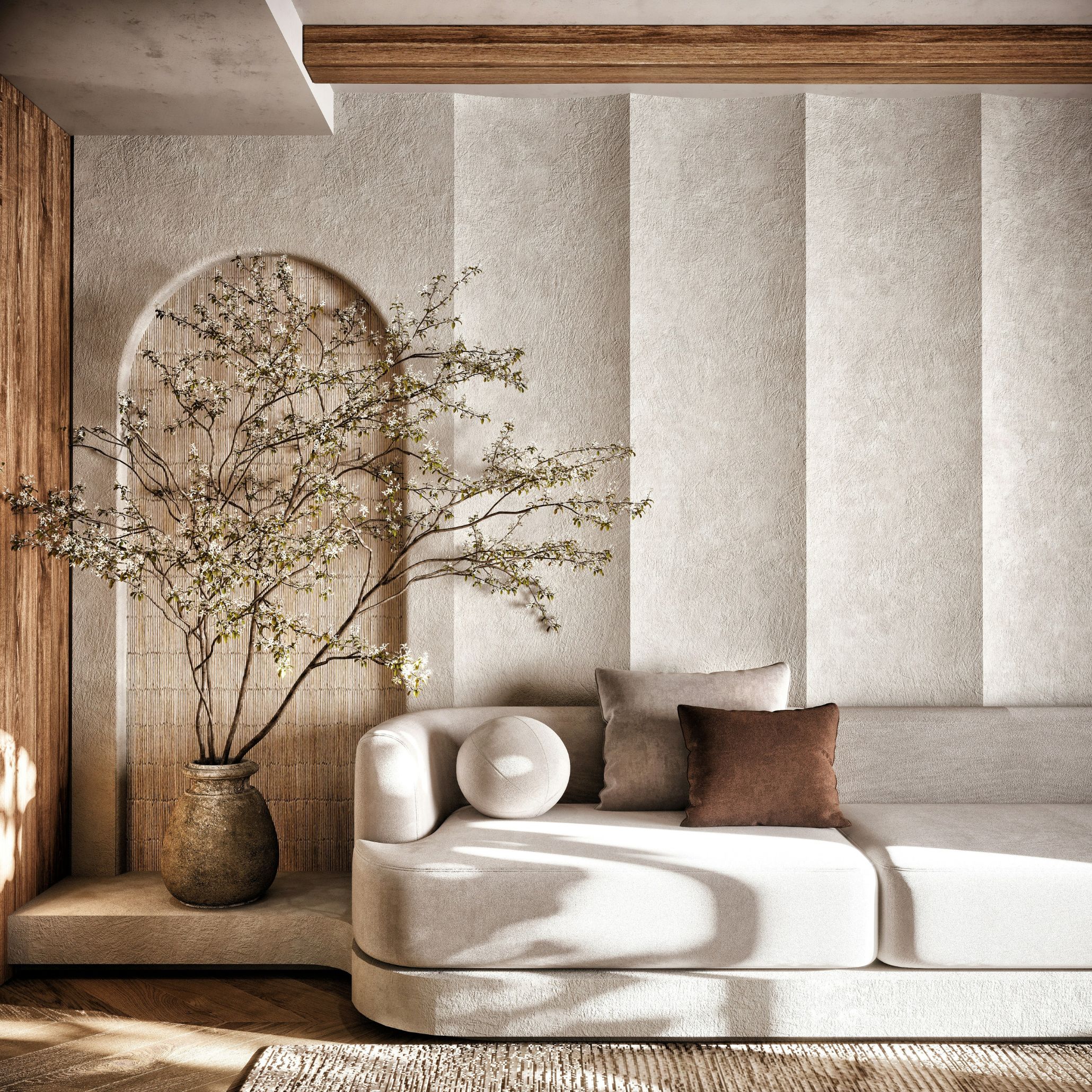 Curved neutral sofa with brown and taupe cushions against a plaster-textured wall, flowering branches in a stone vase, a wellness space shaped through wellness interior design, natural materials and calm proportions.