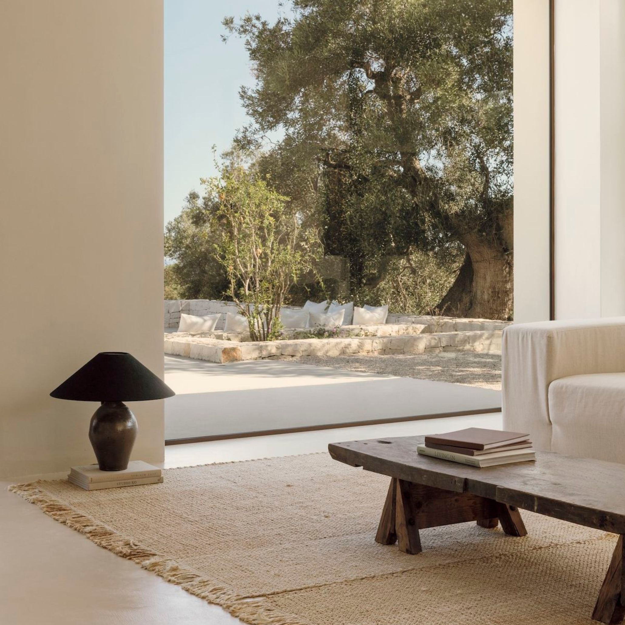 Sunlit living space with linen sofa, dark ceramic lamp and reclaimed wood coffee table opening onto an olive grove terrace, a wellness setting guided by wellness design through natural light and quiet spatial flow.
