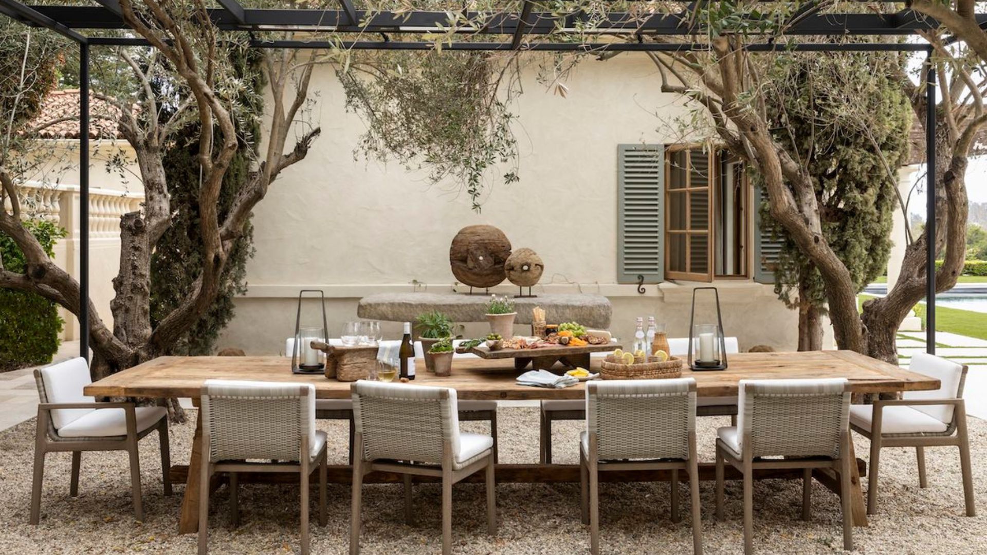 A long wooden table set for an alfresco dining gathering beneath an olive tree pergola, surrounded by woven outdoor furniture chairs and rustic stone accents against a shuttered Mediterranean façade, capturing the leisurely spirit of a summer soirée on the Riviera.