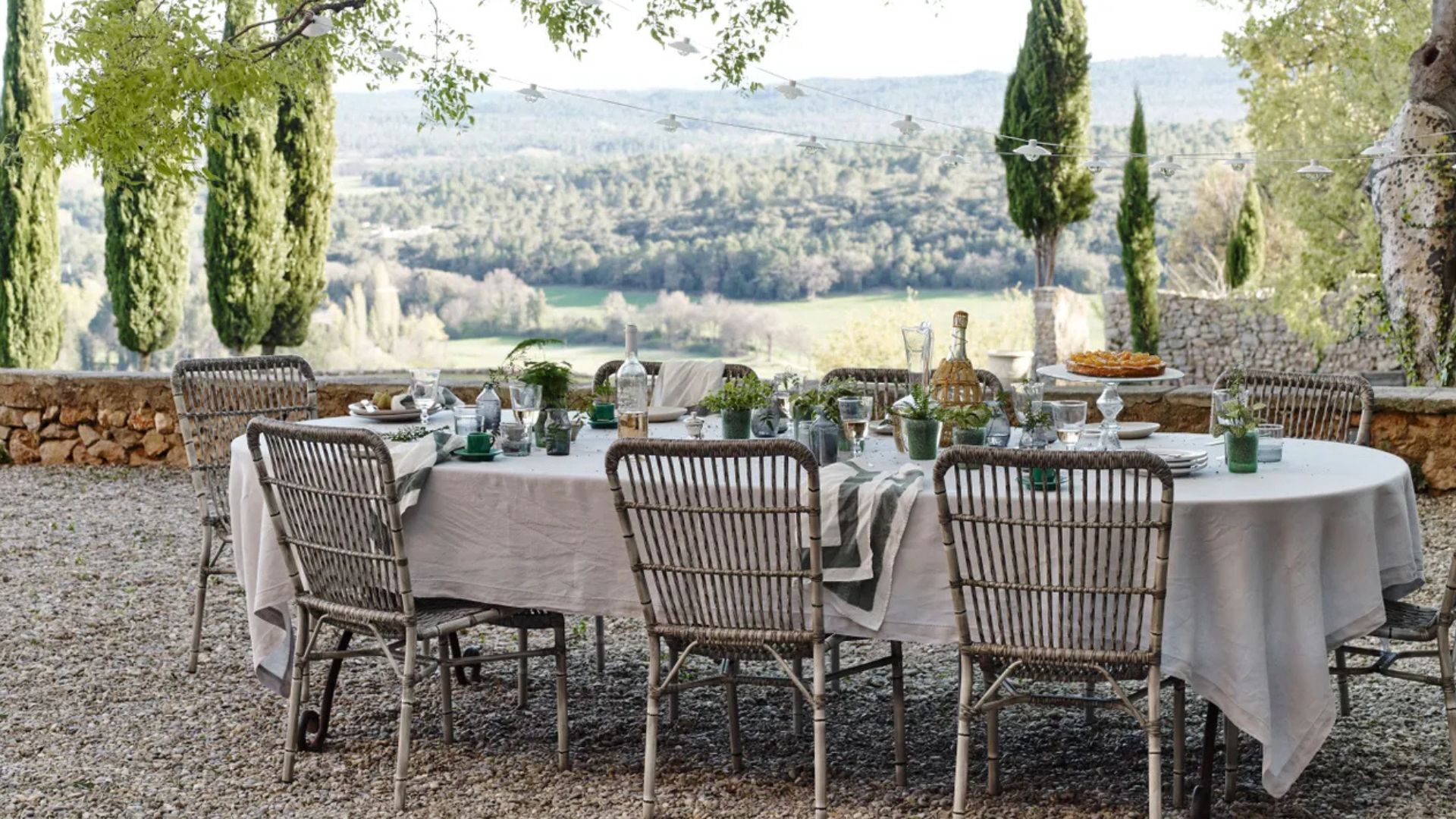A round dining table set with linen and greenery beneath string lights, surrounded by woven rattan chairs on a gravel terrace overlooking rolling cypress-lined hills, an outdoor dining setting shaped by the surrounding Mediterranean landscape.