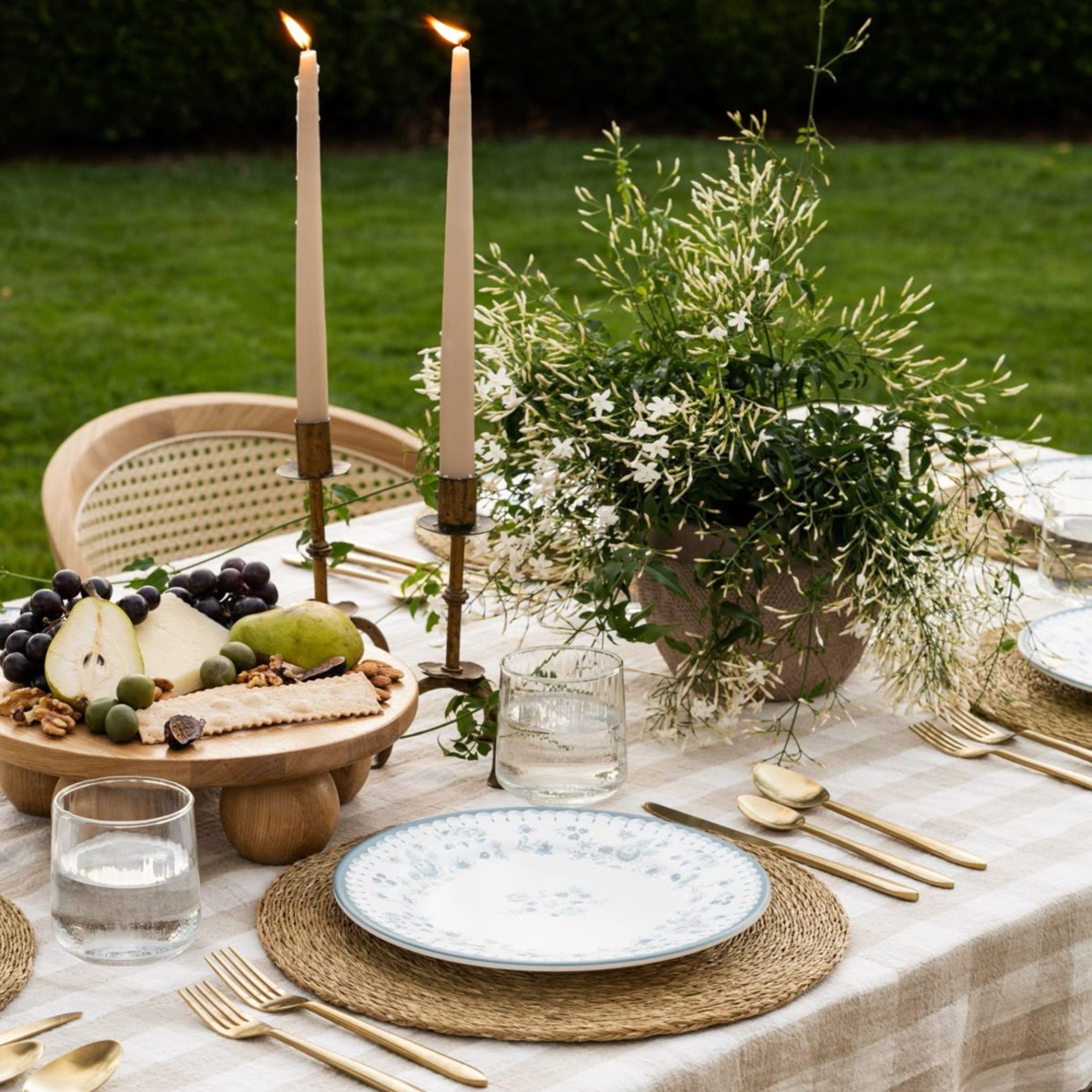 A candlelit garden table set with gold flatware, a wooden cheese board and trailing white jasmine, styled for a relaxed summer soirée on the lawn, an example of alfresco dining table styling.