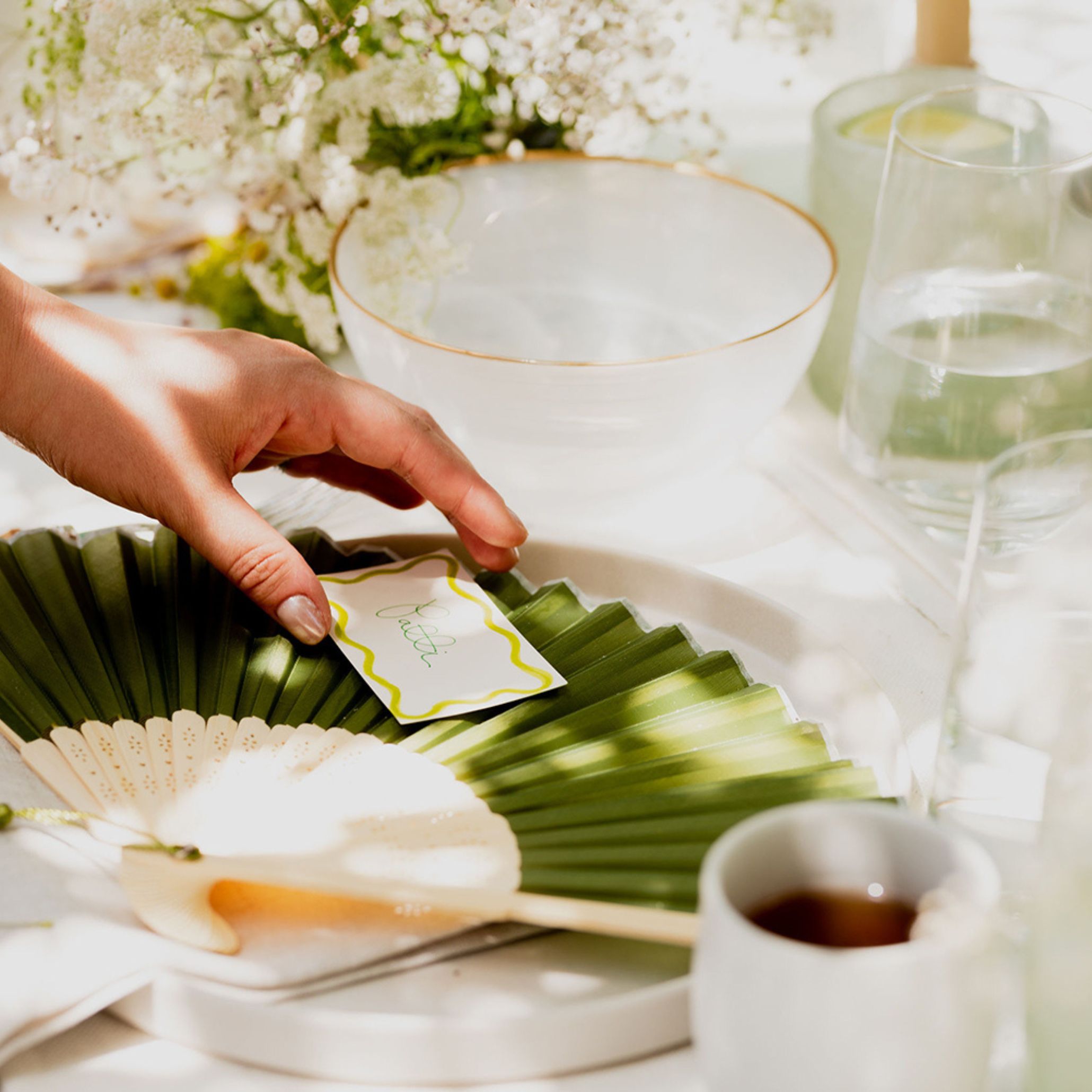 Close-up of a hand placing a handwritten place card on a folded palm-leaf fan atop a white plate, surrounded by gold-rimmed bowls and baby's breath, a delicate detail from an outdoor dining setting.