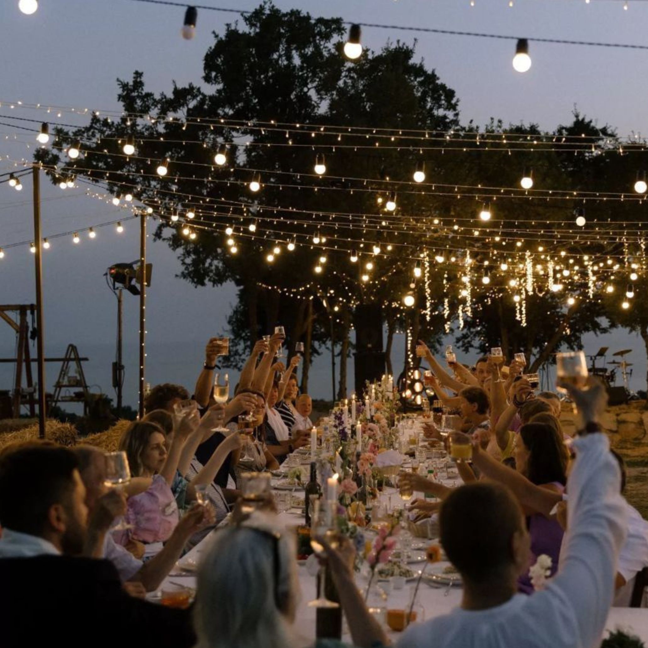 Guests raise their glasses beneath canopies of string lights strung through old trees, gathered around a long candlelit table, capturing the warmth of a Eurosummer evening celebration.