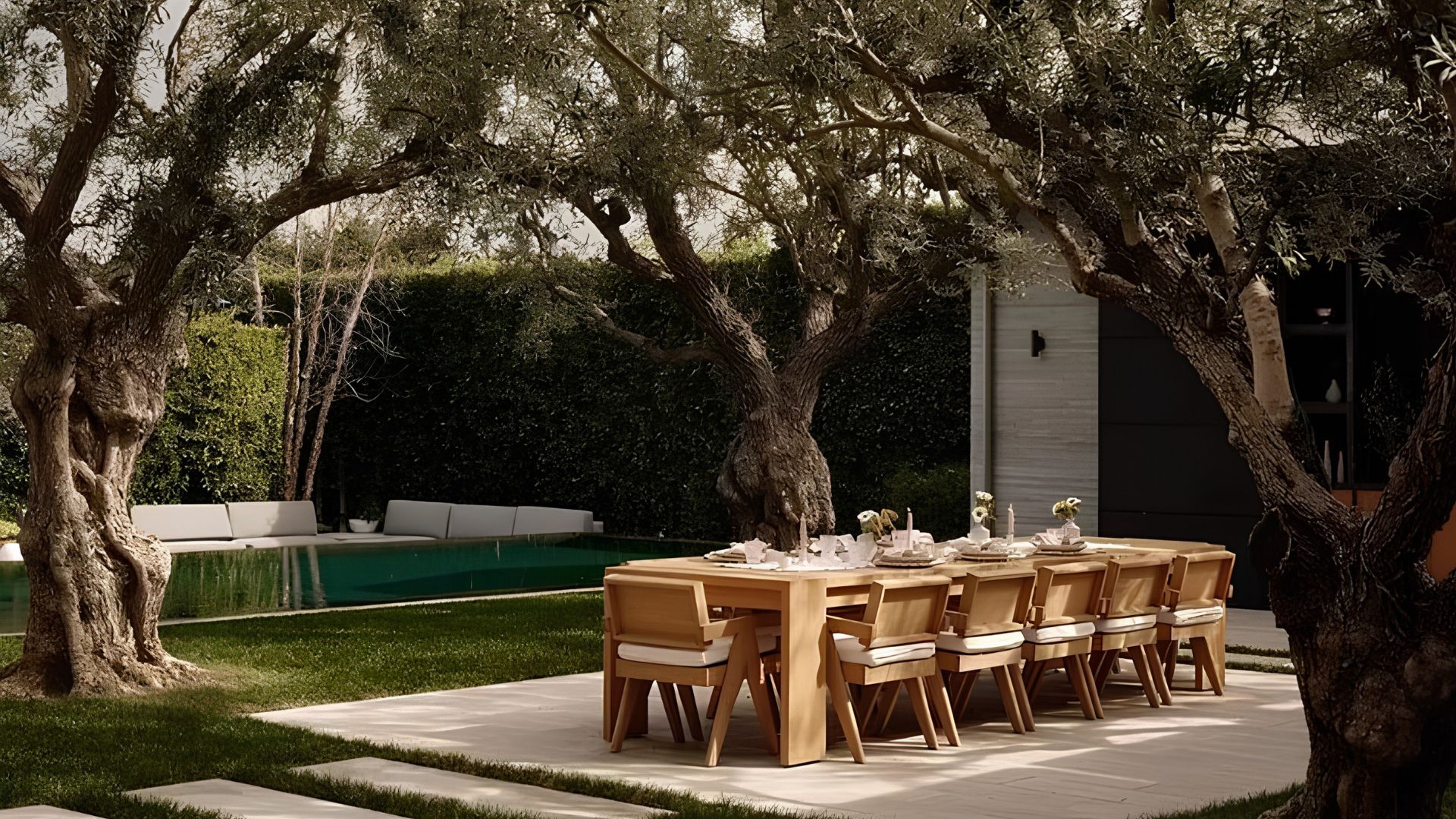 A long teak dining table and chairs set beneath sprawling old olive trees beside a pool and lounge, capturing the effortless spirit of an alfresco dining area designed for lingering summer meals.