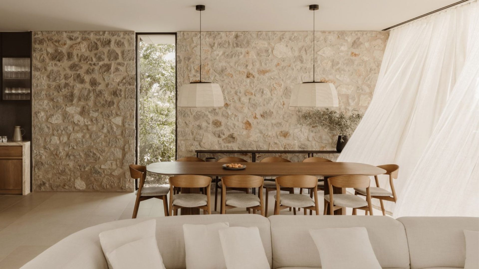 An open-plan dining and living area featuring a raw stone accent wall, oval wooden dining table with woven-back chairs, and linen drapery framing garden views, an example of luxury dining room furniture paired with contemporary furniture in a rustic greek setting true to Mediterranean Interior design.