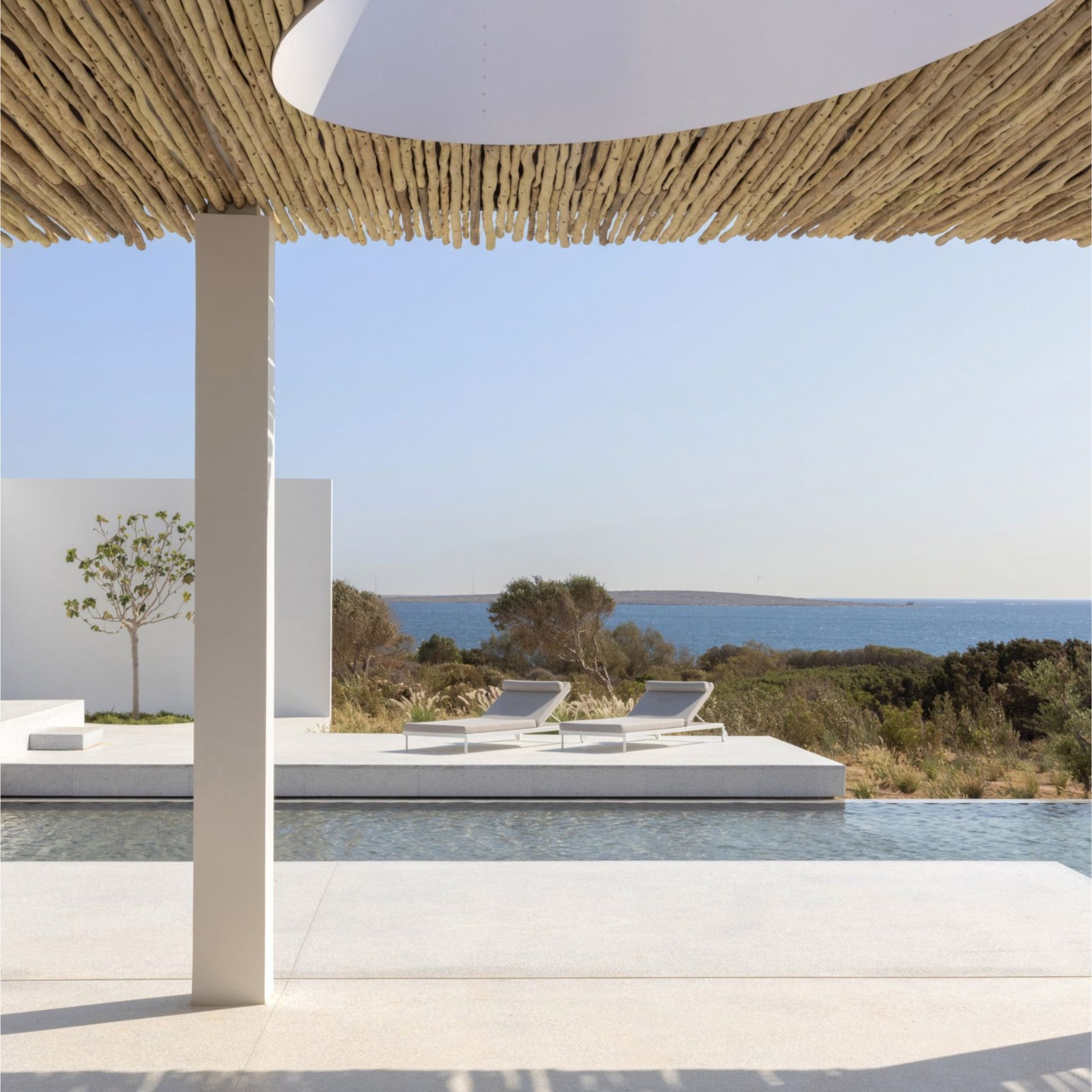 A poolside view framed by a driftwood-covered pergola, with two white sun loungers set against wild coastal greenery and open sea, an example of minimalist design and luxury furniture suited to a wellness center furniture design setting.