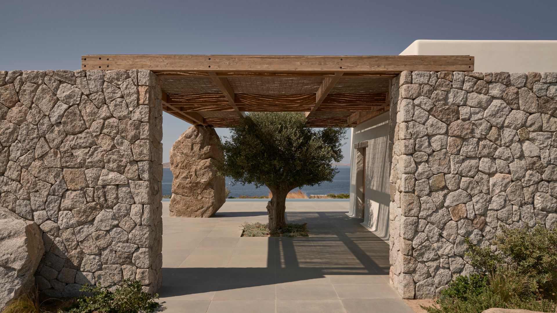 A stone-walled entrance framing a mature olive tree and distant sea view, topped with a timber and reed pergola roof, showing the raw wood texture and craftsmanship central to rustic greek architecture and Mediterranean Interior design.