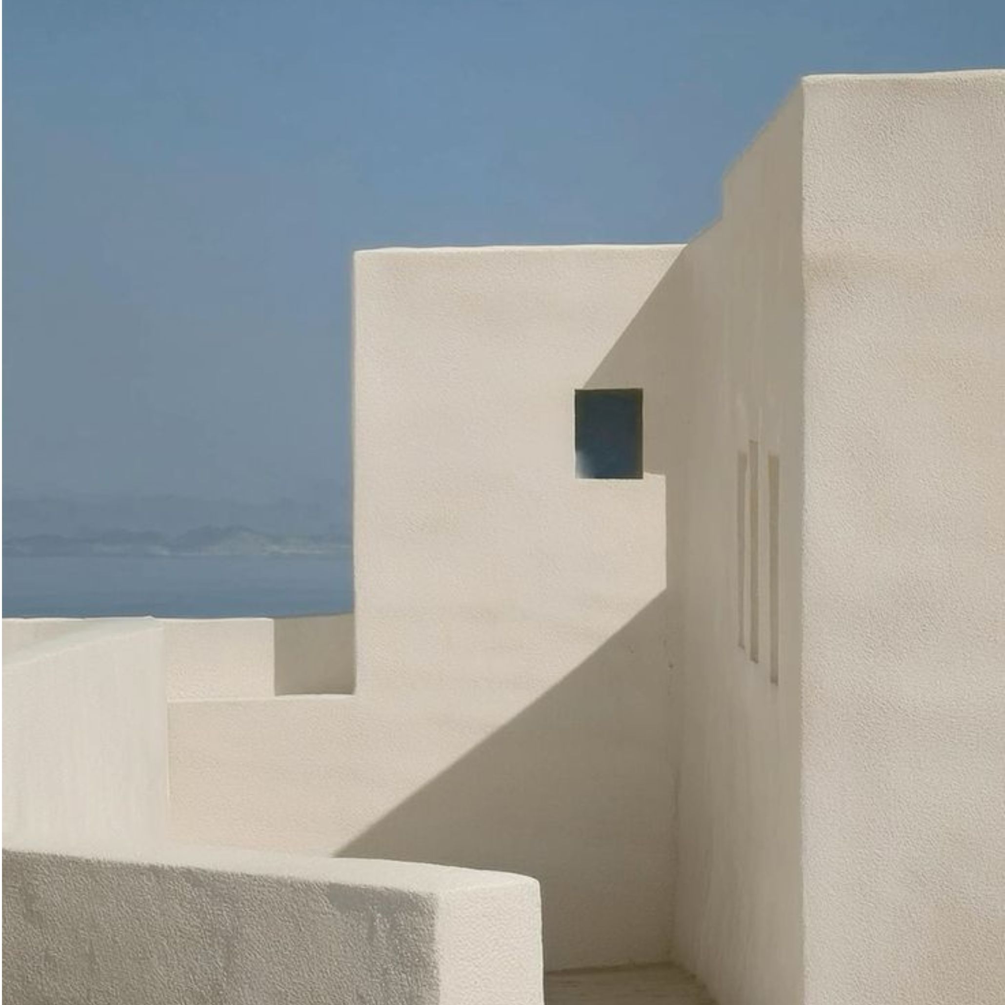 Sculptural whitewashed exterior walls with sharp geometric shadows and a single dark blue window against the sea and sky, illustrating the minimalist design and pure architectural form central to rustic greek and Mediterranean Interior design.