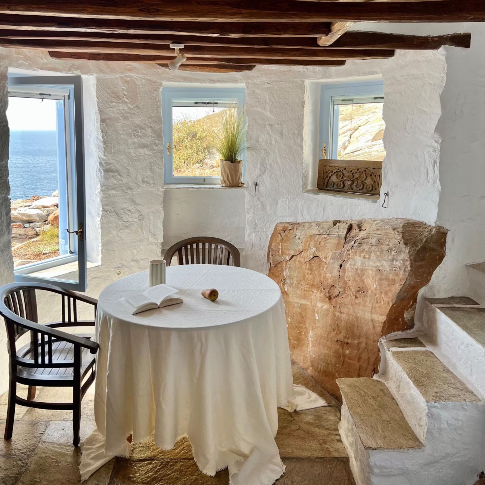 A stone-walled interior with a linen-draped round table, wooden armchairs and a rustic exposed timber beam ceiling, sea views through open windows, an example of bespoke furniture and handmade furniture true to rustic greek character.