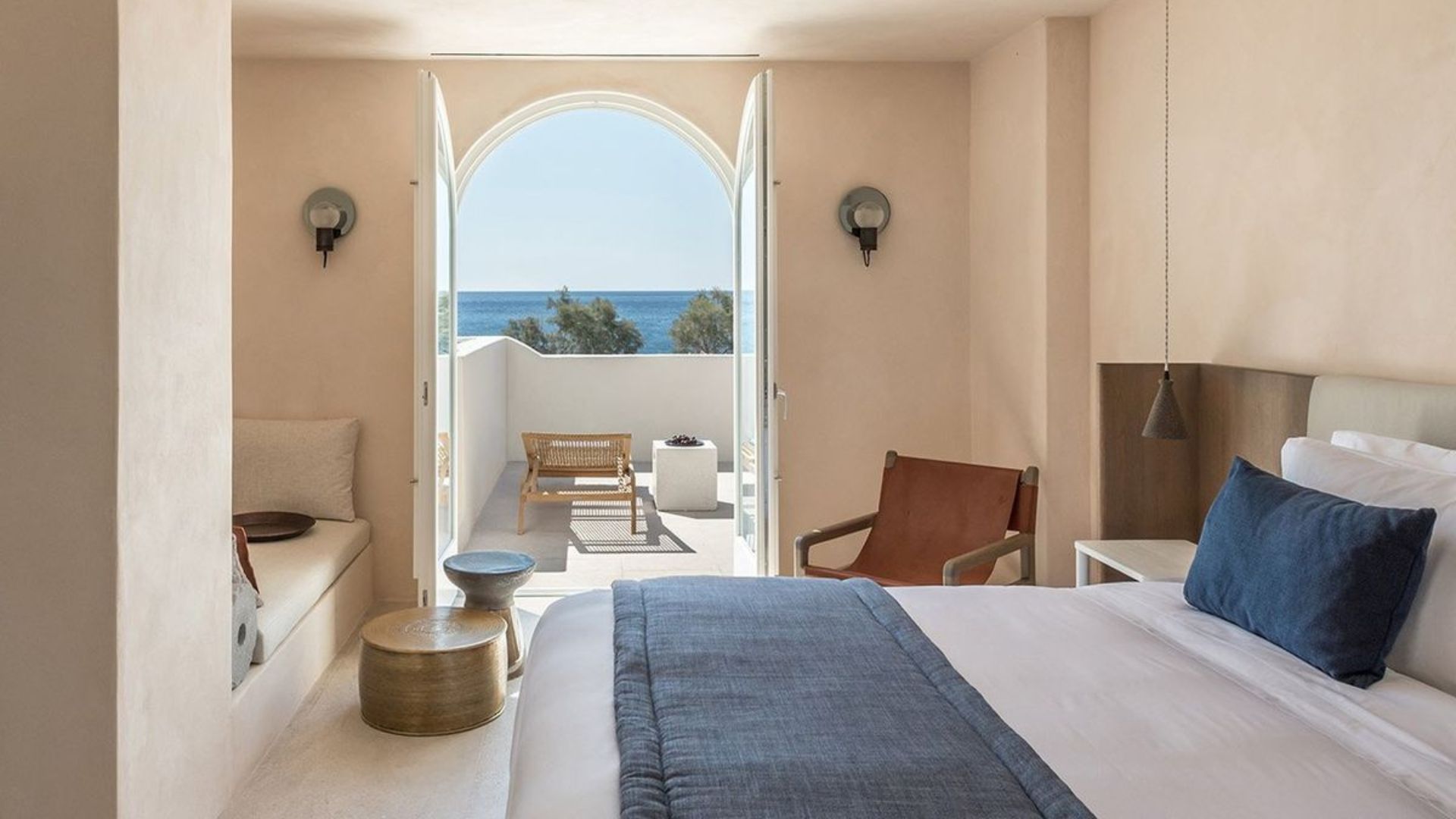 A sand-toned bedroom with a leather armchair and blue-linen dressed bed, opening through an arched doorway to a sea-view terrace, showing the warm texture and luxury bedroom furniture palette that defines rustic greek and Mediterranean Interior design.